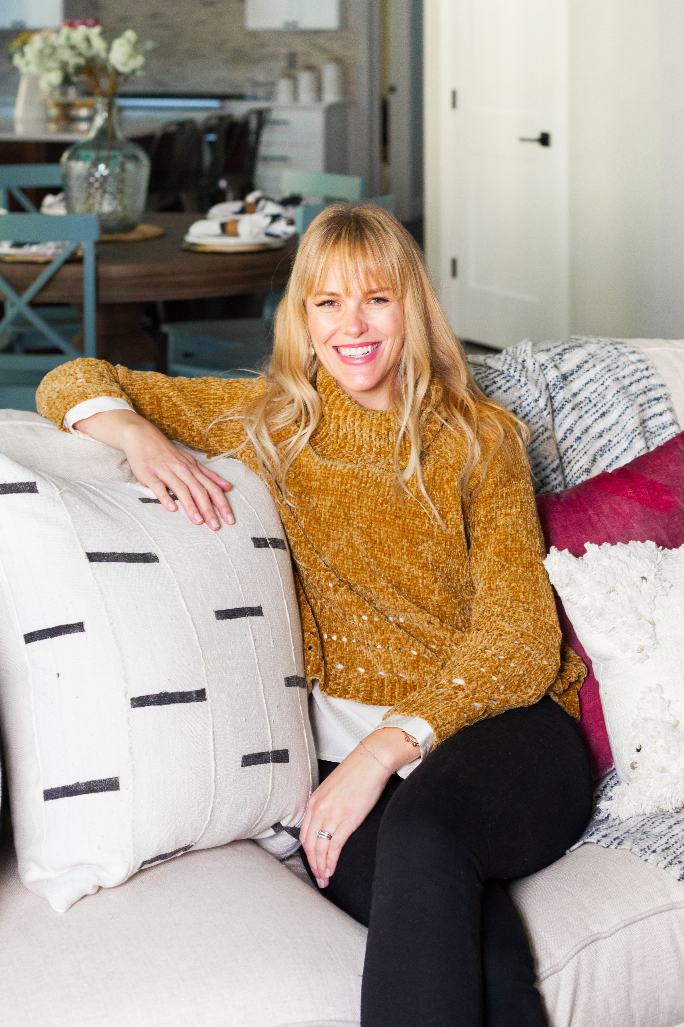 A woman sitting on a beige sofa, smiling, with her left arm resting on a large cream pillow with black lines. She has blonde hair with bangs and is wearing a mustard-colored sweater and black pants. Behind her are decorative pillows, and in the background, a dining table with a large glass vase of white flowers and a blurred kitchen area.