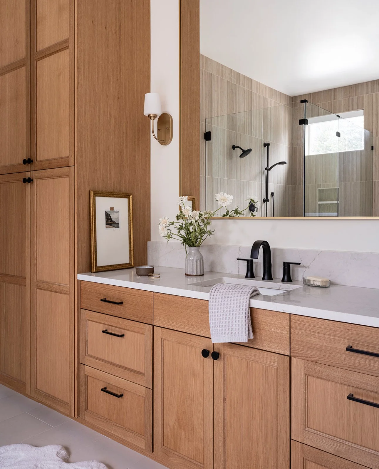 Modern bathroom with wooden cabinetry, black fixtures, and a marble countertop with a towel, soap, and a vase with flowers. A framed photo and a wall sconce are on the wall, and a large mirror reflects a walk-in shower with beige tiles and black fixtures.