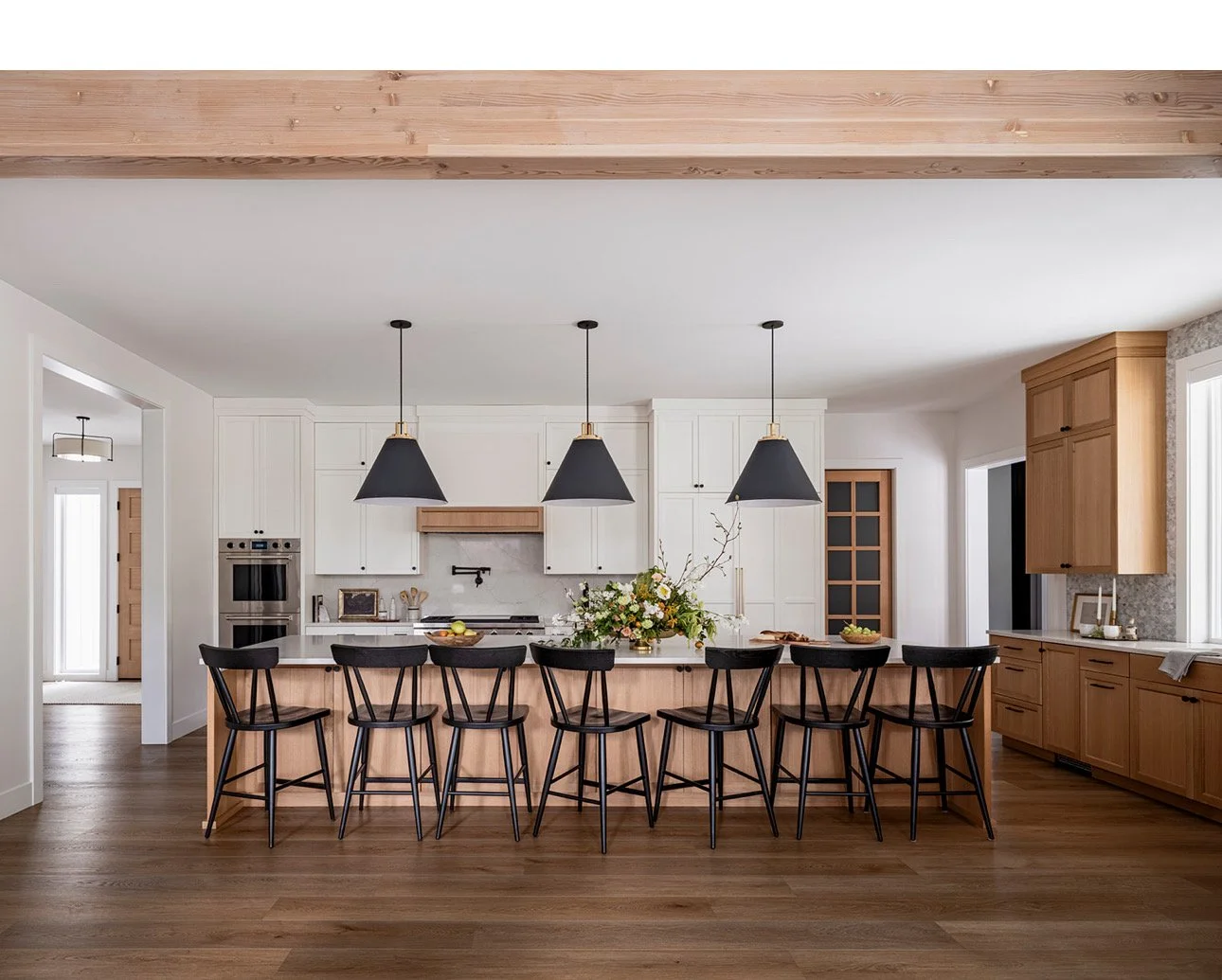 Modern kitchen with a large island, eight black chairs, and pendant lights, featuring white cabinets, wooden accents, and a dark hardwood floor.
