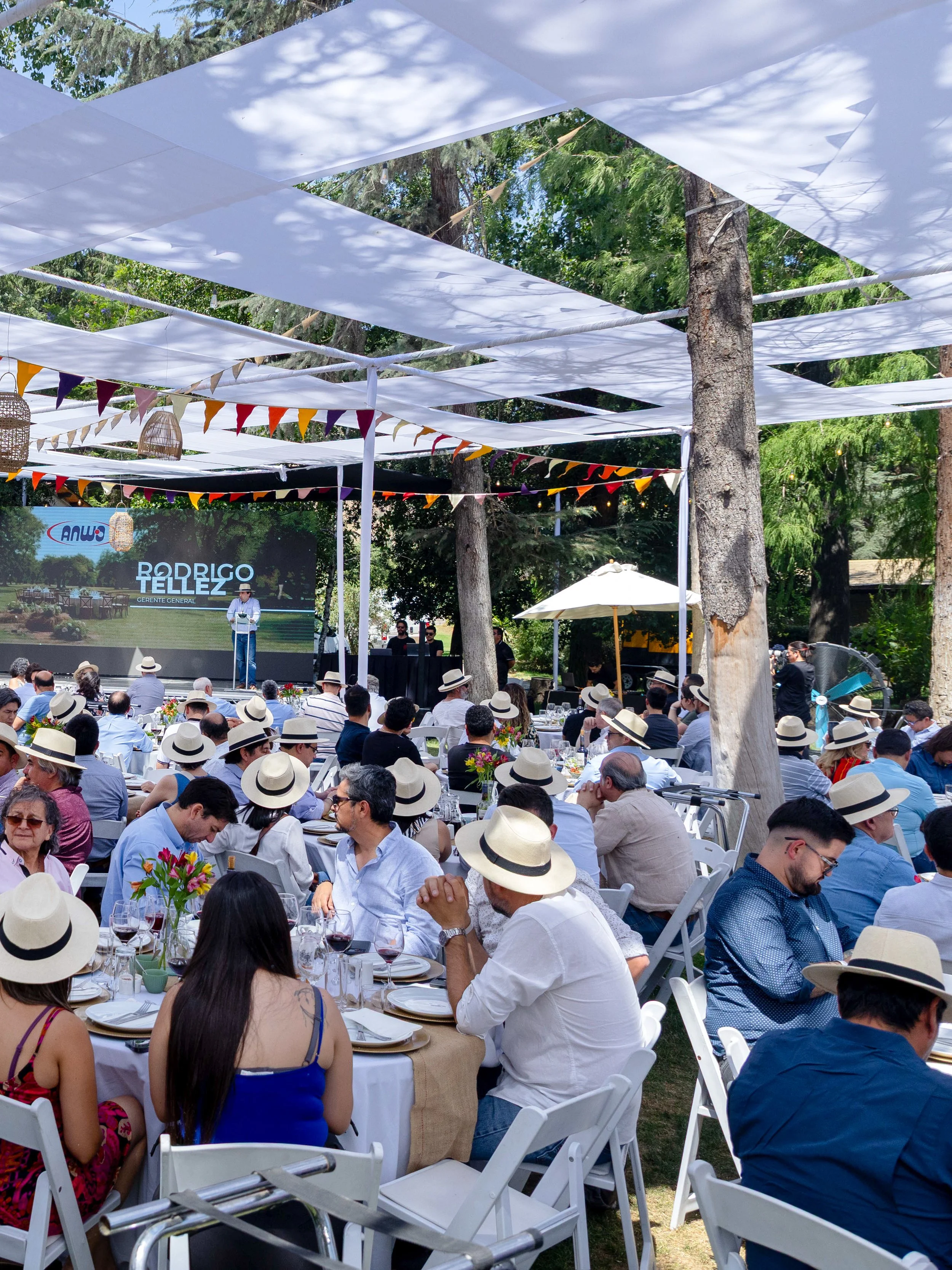 An outdoor event with many people seated at tables, wearing straw hats. There is a stage with a person speaking into a microphone, and the backdrop displays the name Rodrigo Tellez as the general manager. Decorations include white fabric canopy and colorful triangular flags. The setting is surrounded by trees.
