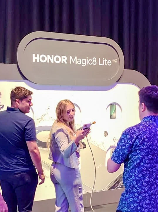 Three people standing in front of a large display advertising the HONOR Magic8 Lite smartphone at an event. One woman is holding a smartphone and appears to be demonstrating or explaining something to two men.