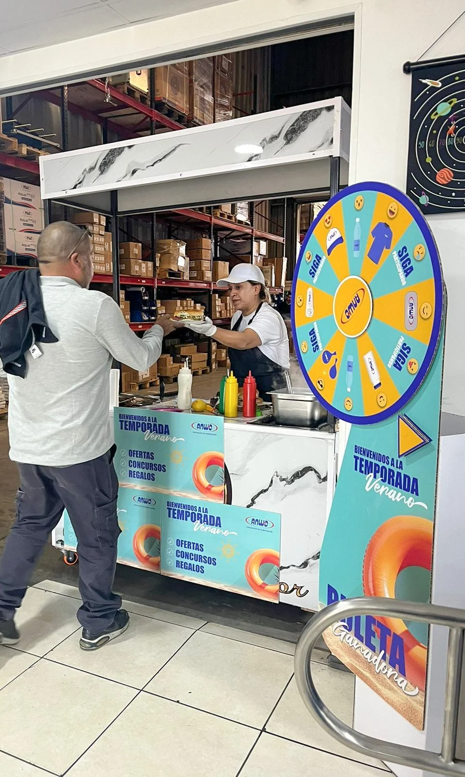 A man orders food from a vendor at a promotional booth with a spinning wheel for prizes. The booth is set up in a warehouse or storage area with shelves of boxes in the background. The vendor is handing a plate of food to the man, who is wearing a white jacket and dark pants.