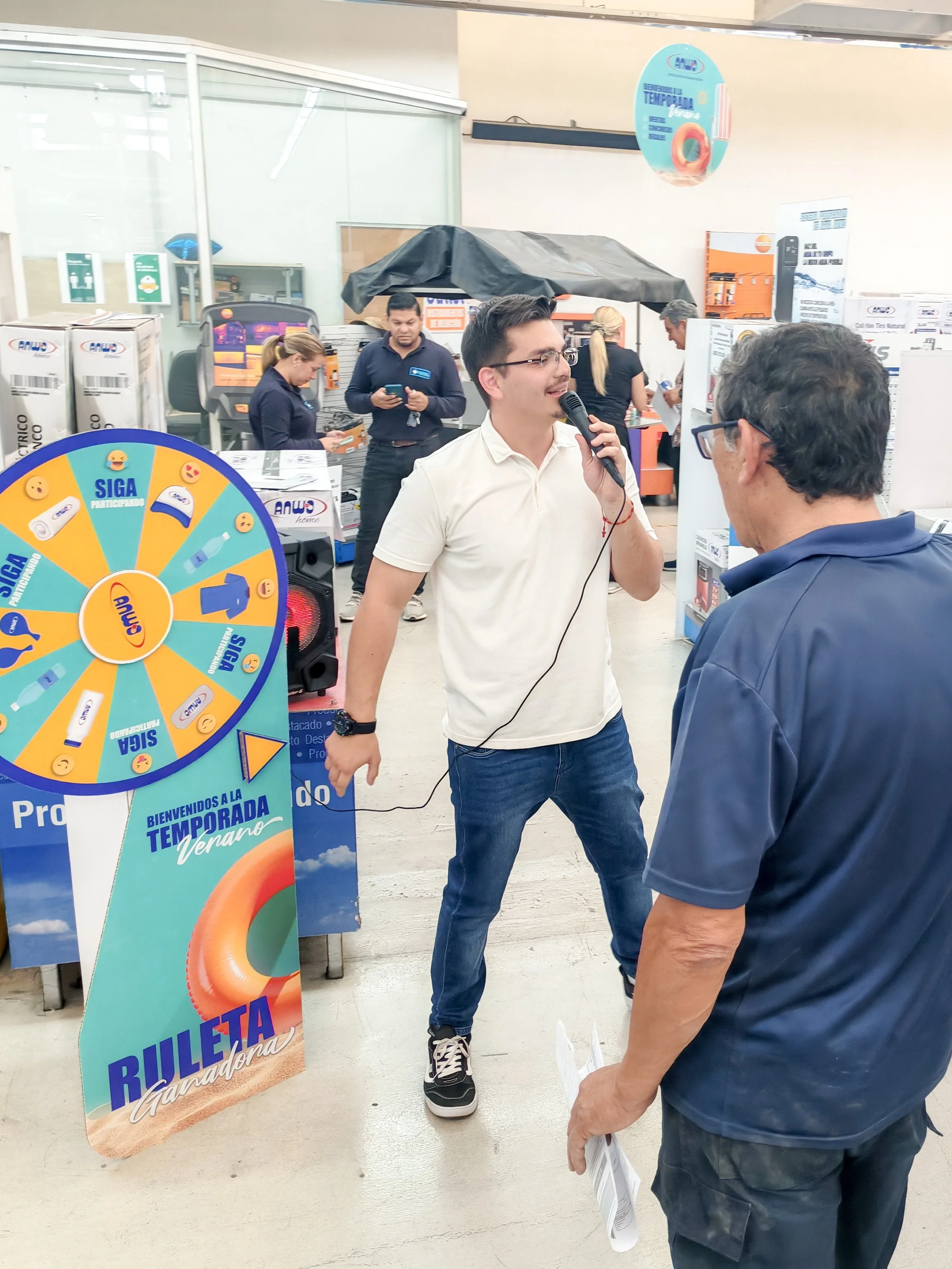 A young man with glasses, wearing a white shirt, holds a microphone and talks to an older man wearing glasses and a navy shirt, at a promotional event inside a store. There are posters, a spinning prize wheel, and store staff in the background.