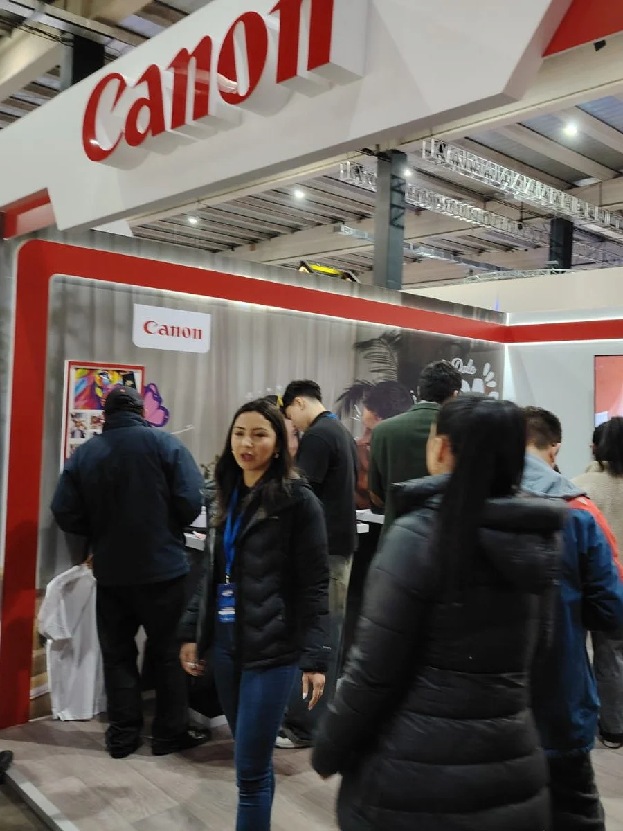 People at a Canon booth at a trade show or exhibition, with the Canon logo displayed prominently overhead.