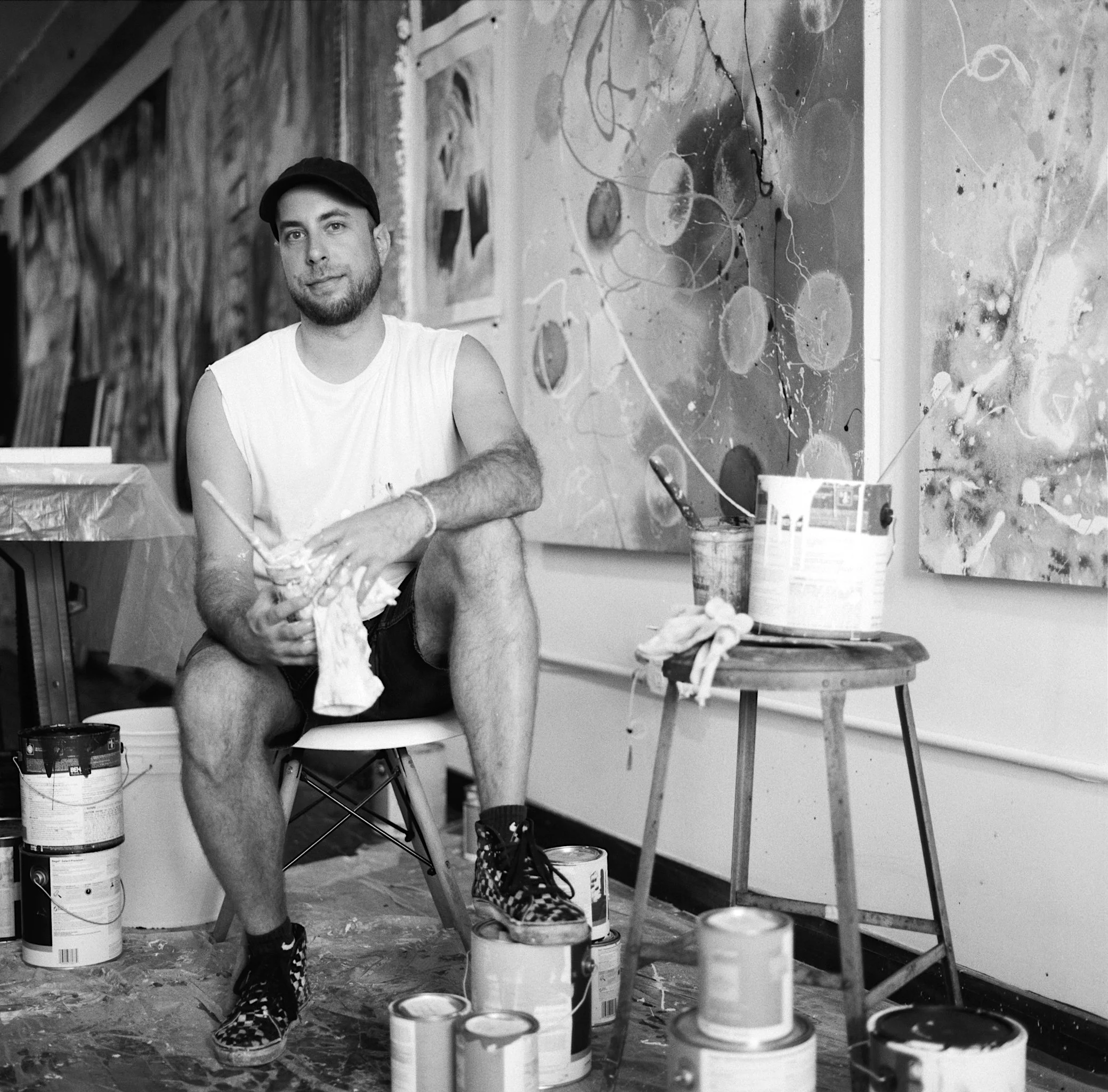 A man wearing a cap, sleeveless shirt, and checkered shoes sitting on a chair in front of painted walls, surrounded by paint cans and art supplies, suggesting an art studio.