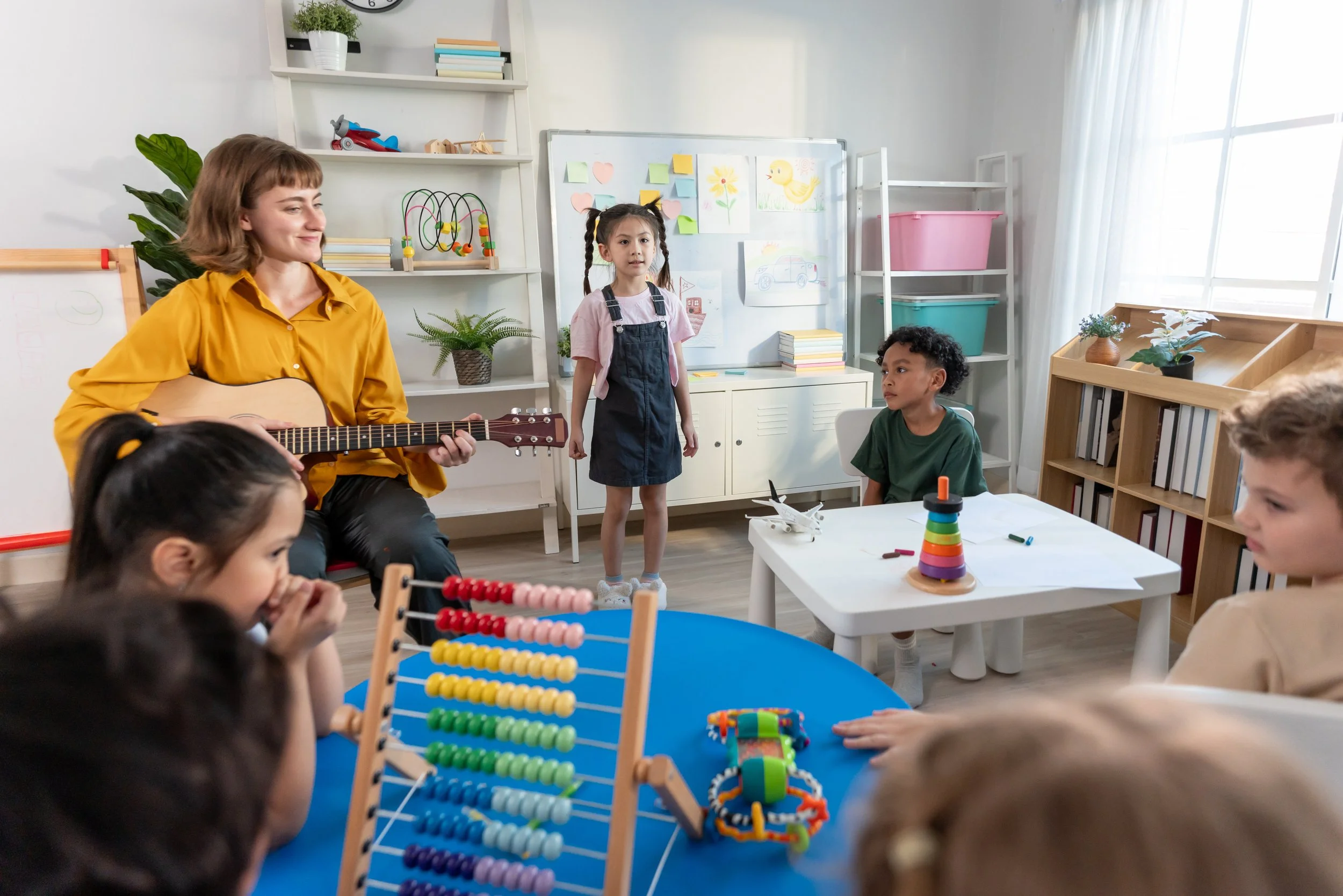 A woman playing guitar in a classroom with children. Two children stand nearby with a whiteboard decorated with drawings and notes in the background. The classroom has books, plants, toys, and colorful educational materials.