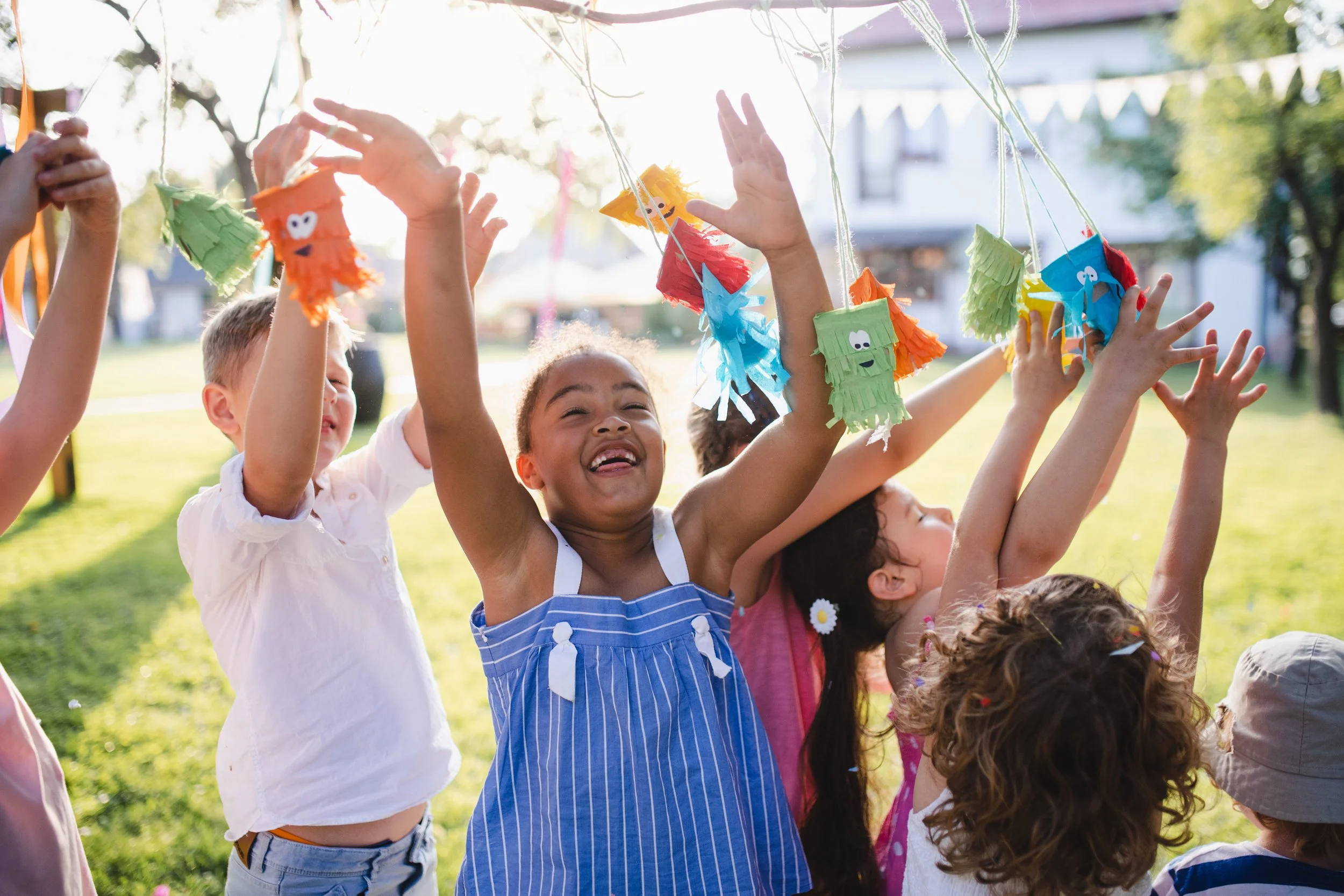 Children are playing outside and reaching up to colorful piñatas hanging from a clothesline, with sunlight and trees in the background.