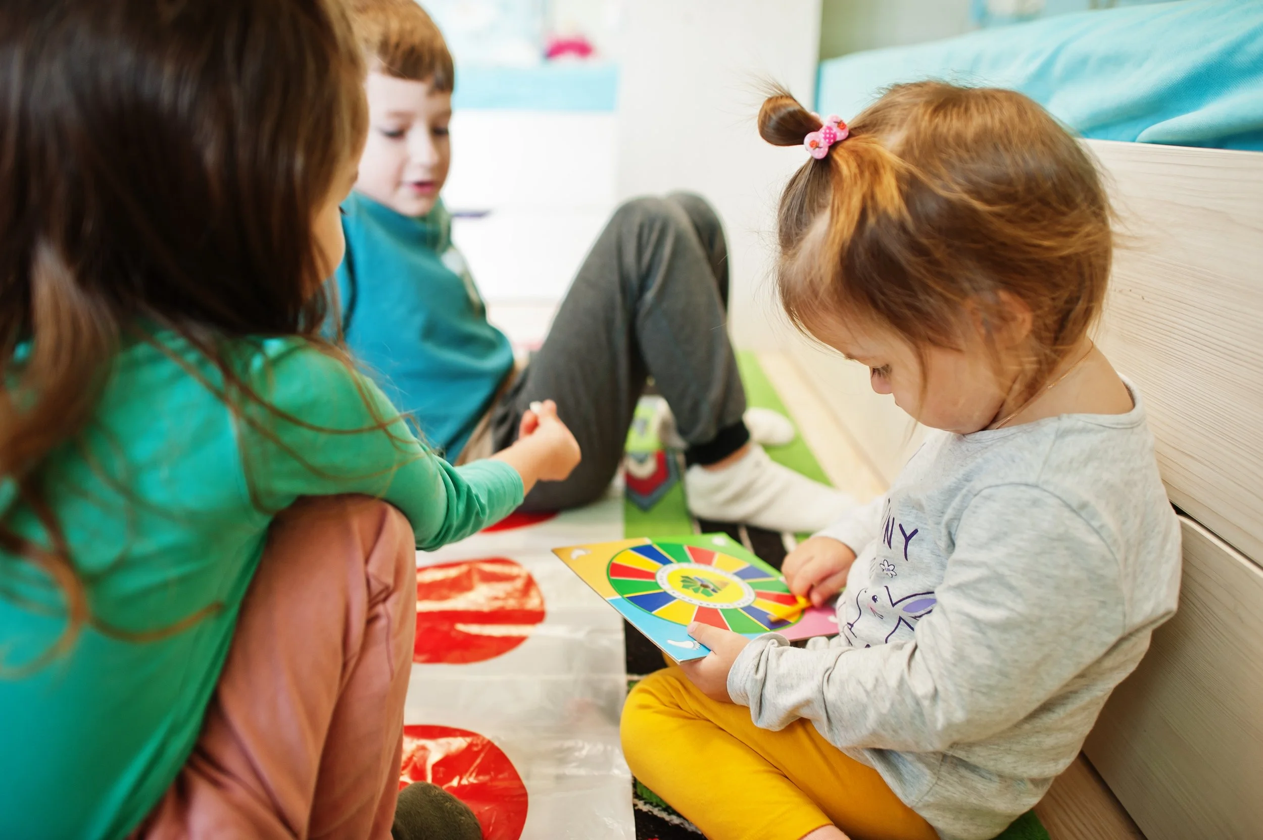 Three children, two boys and one girl, sitting on the floor, playing a colorful board game together in a room with a bed and a wall with a picture and poster in the background.