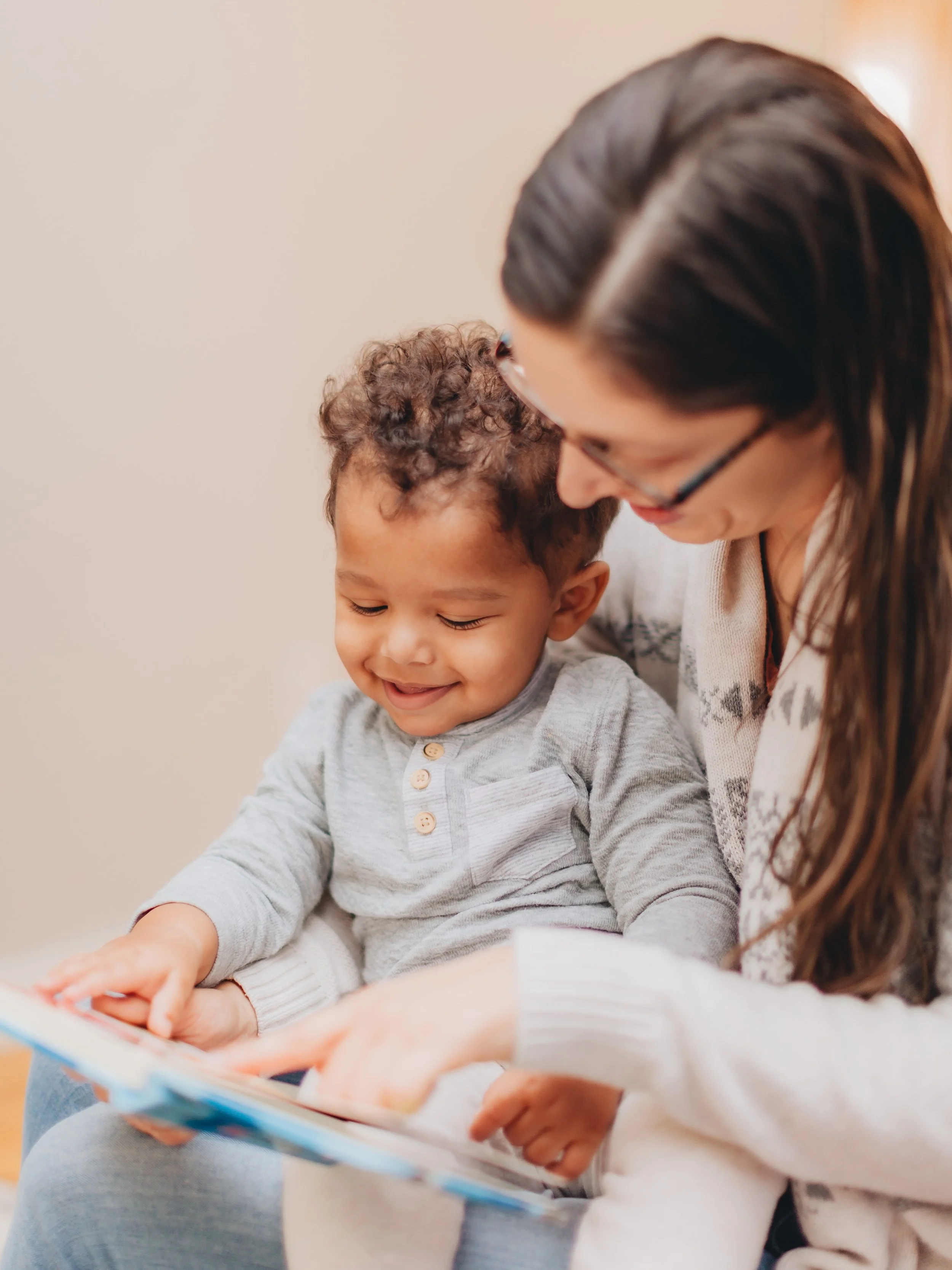 A woman with long brown hair and glasses is leaning in to a young boy with curly hair, sitting closely together, looking at a book. The boy is smiling and engaged, wearing a light gray shirt with buttons, and the woman is wearing a patterned sweater.