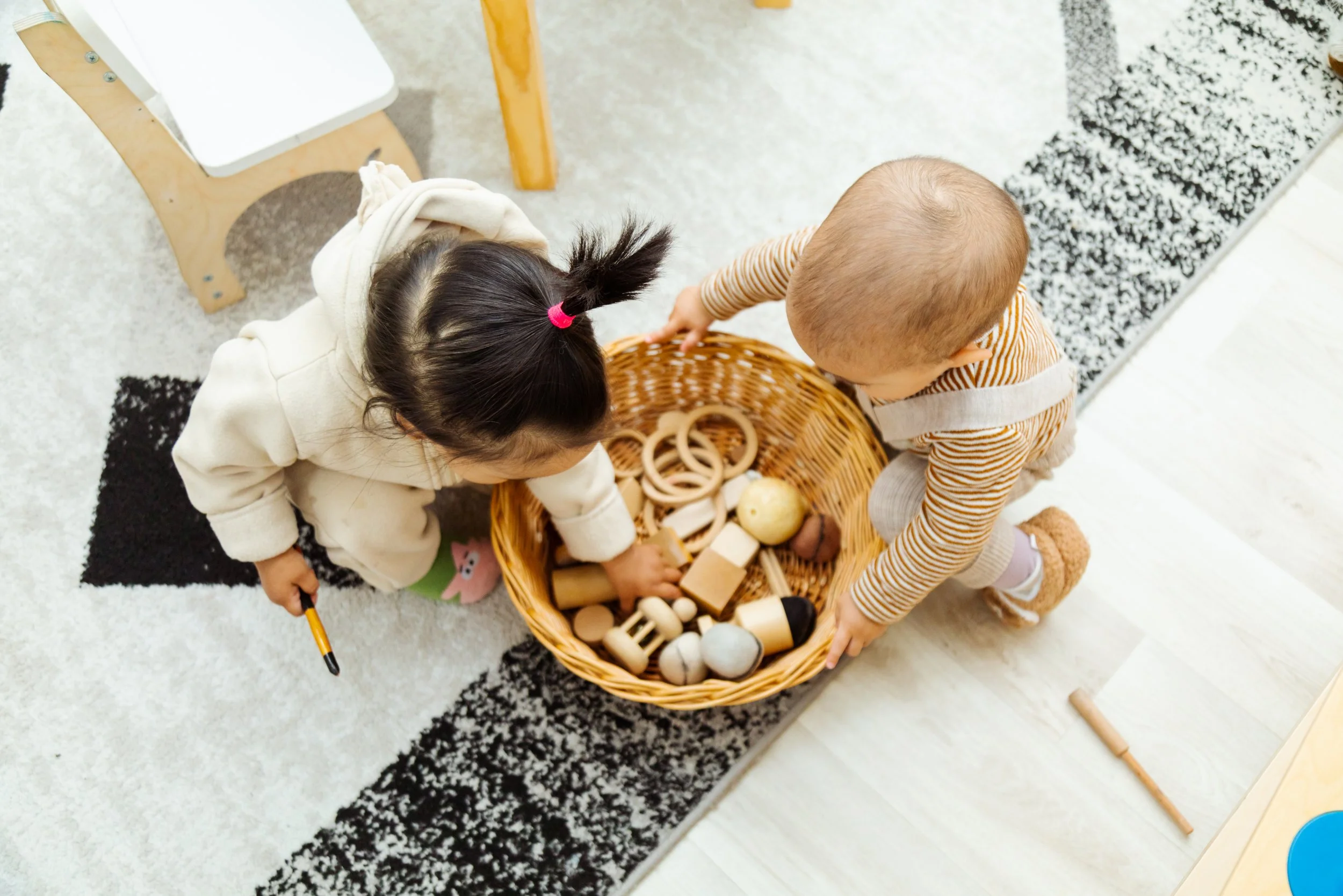 Two children playing with a basket of wooden toys on a black and white rug indoors.