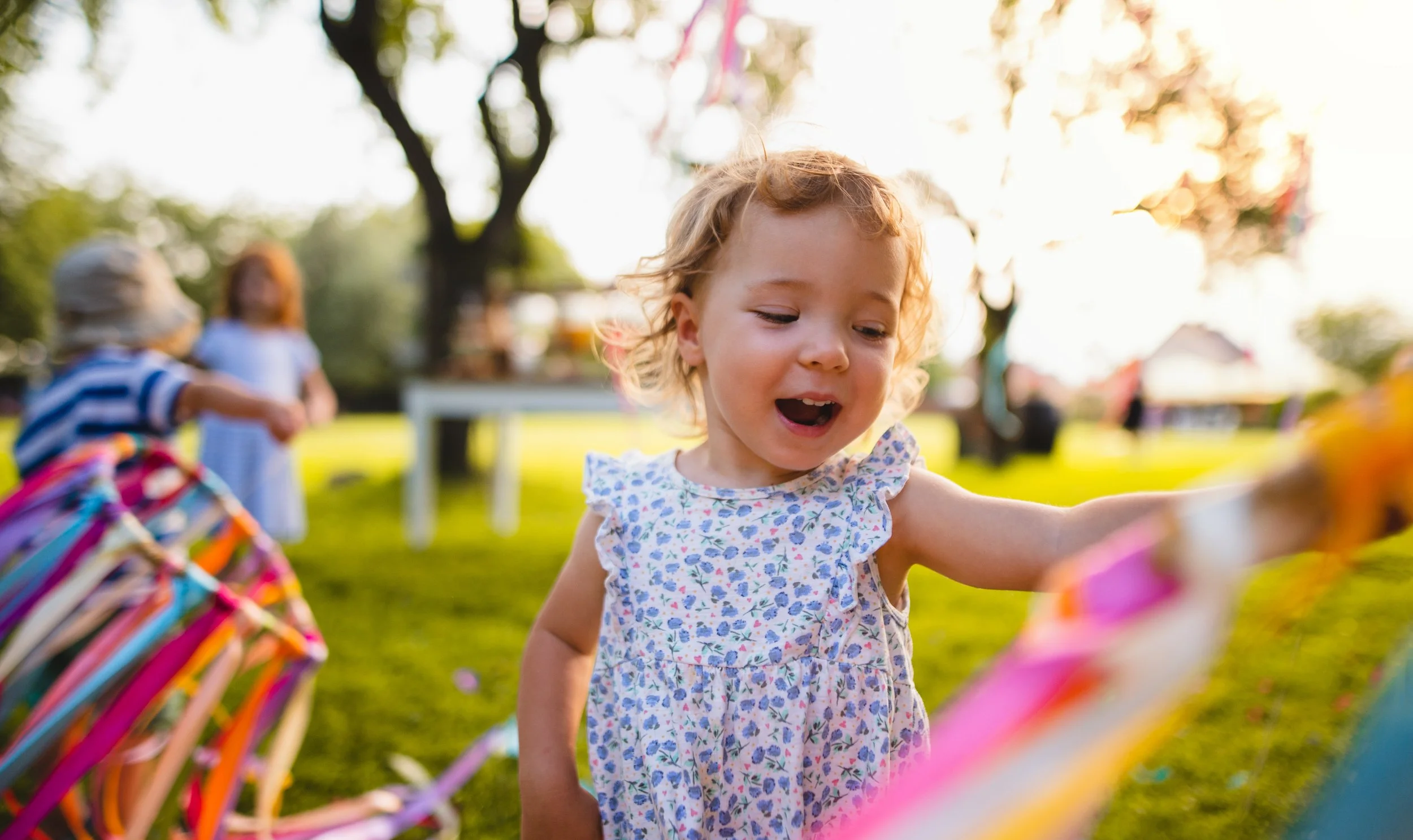 A young girl with curly red hair wearing a floral dress, happily playing outdoors during the daytime with colorful decorations in the background.
