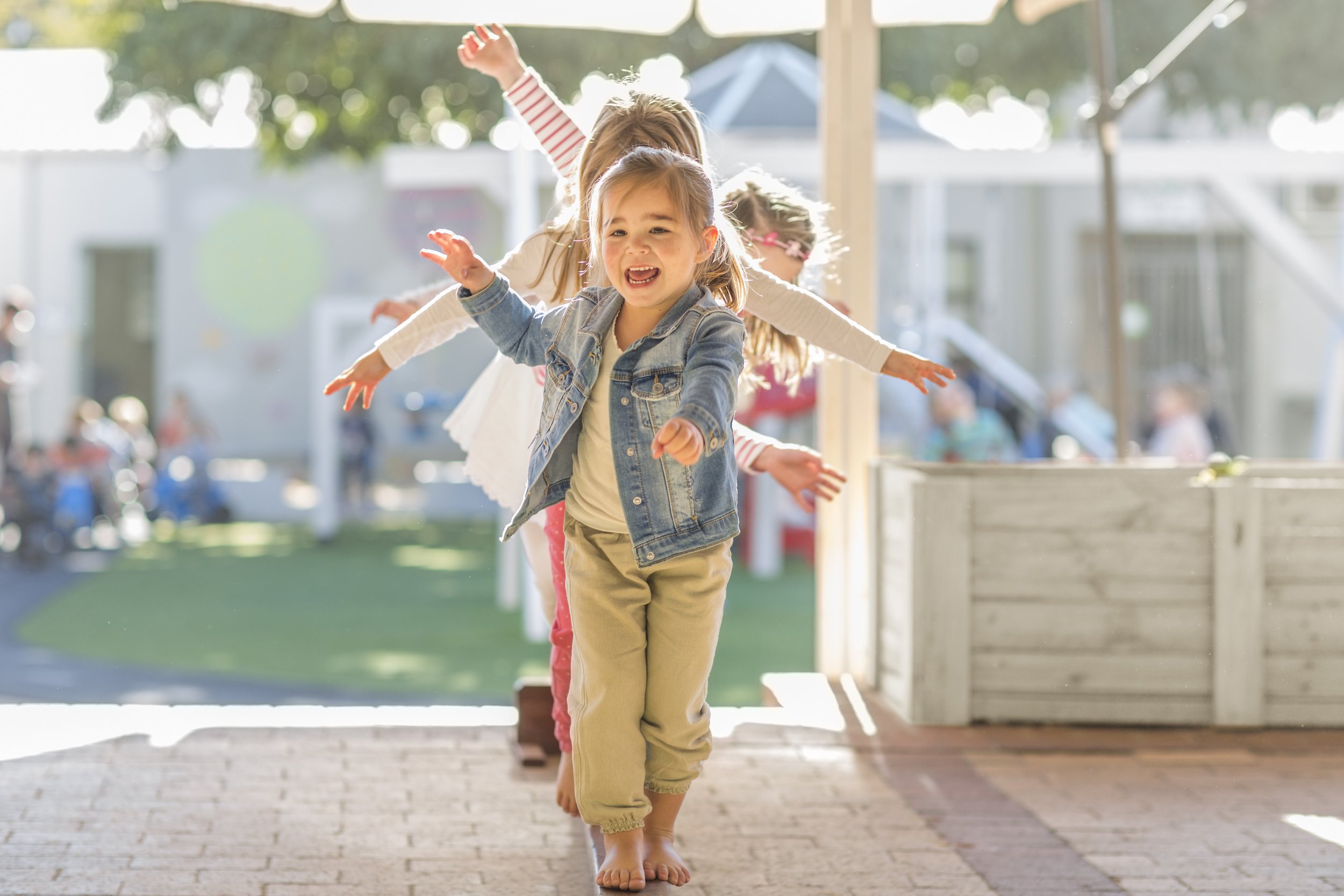 Three young girls are enjoying a sunny day outdoors, walking barefoot through a patio area. The girl in the front is wearing a denim jacket and khaki pants, with her arms outstretched and a big smile. The other two girls behind her are also smiling and have their arms extended, suggesting they are playing or dancing.