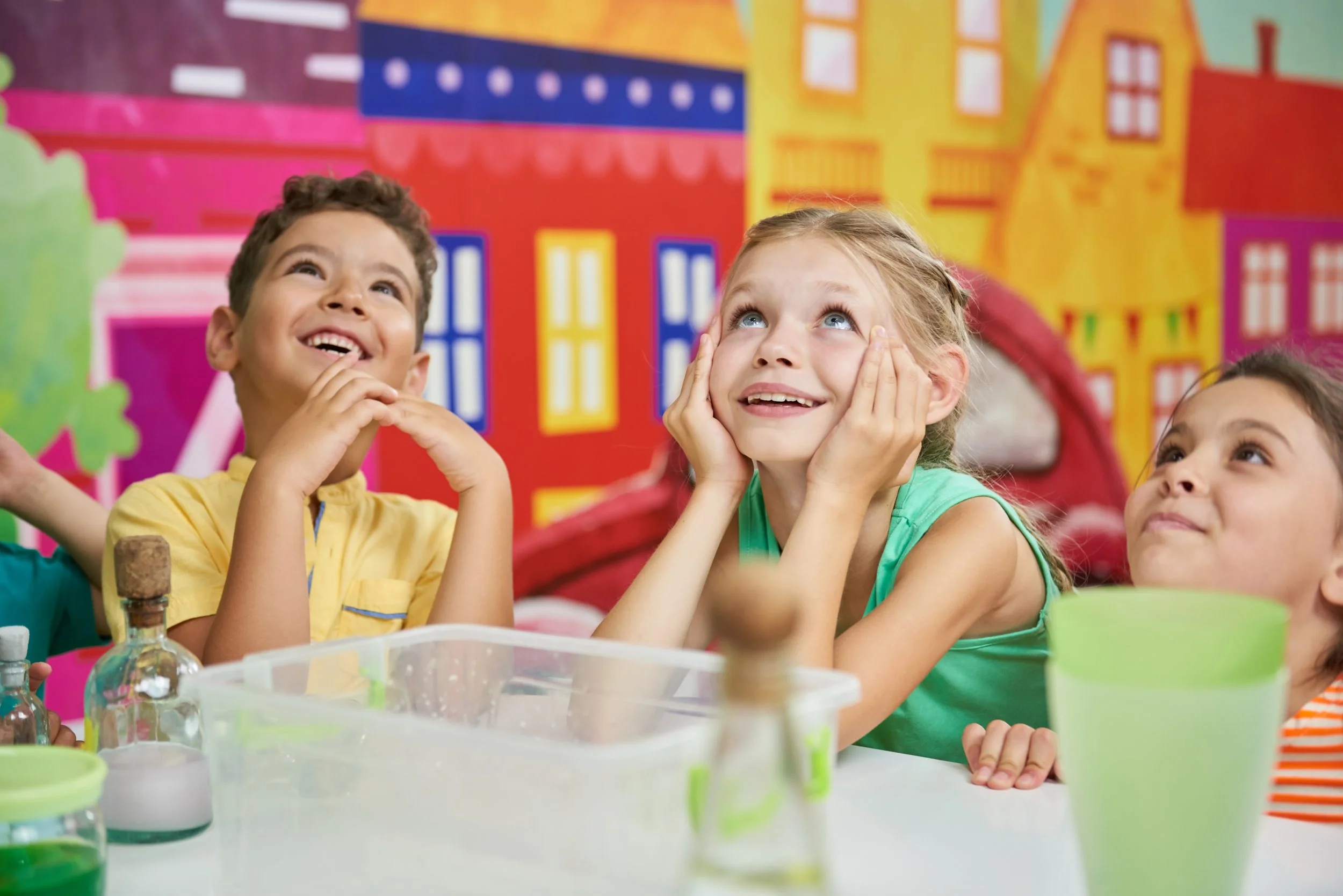 Two children, a boy and a girl, sitting at a table with science experiments, smiling and looking upwards. Bright, colorful background with painted houses.
