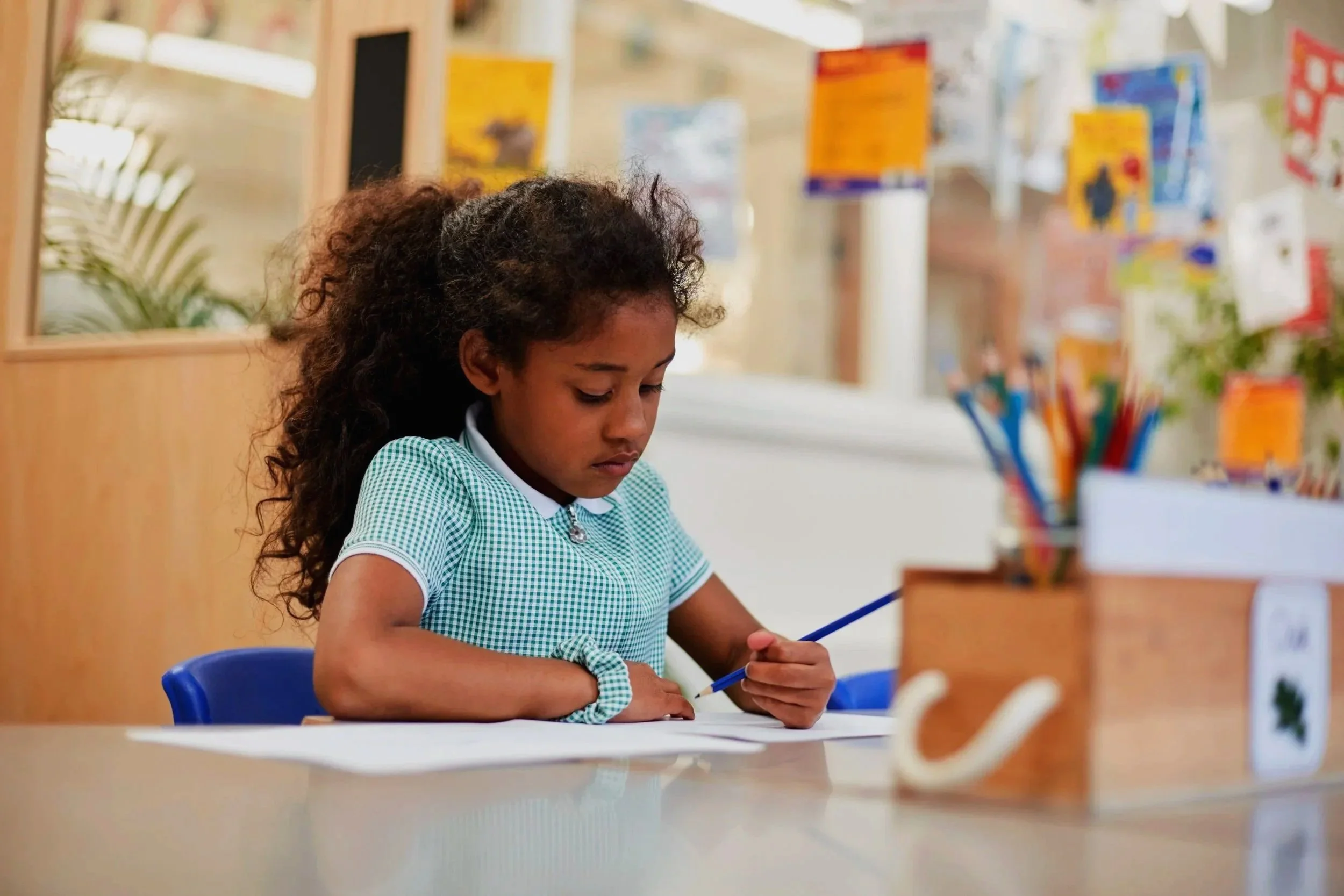 A young girl with dark curly hair sitting at a desk in a classroom, writing or drawing with a blue pencil, wearing a green and white checkered school uniform.