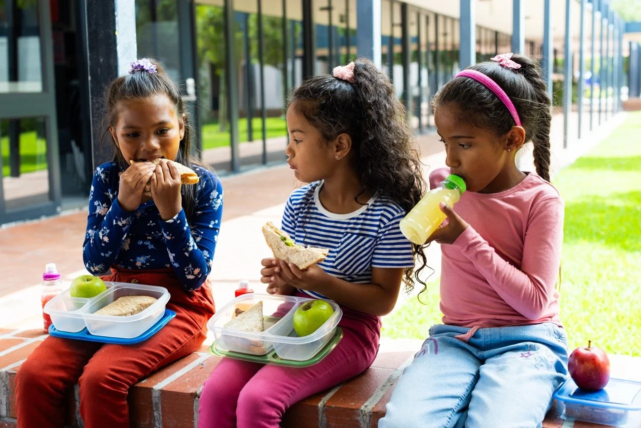 Three young girls sitting outdoors on a brick ledge eating lunch with lunch boxes, sandwiches, apples, and a bottle of juice.