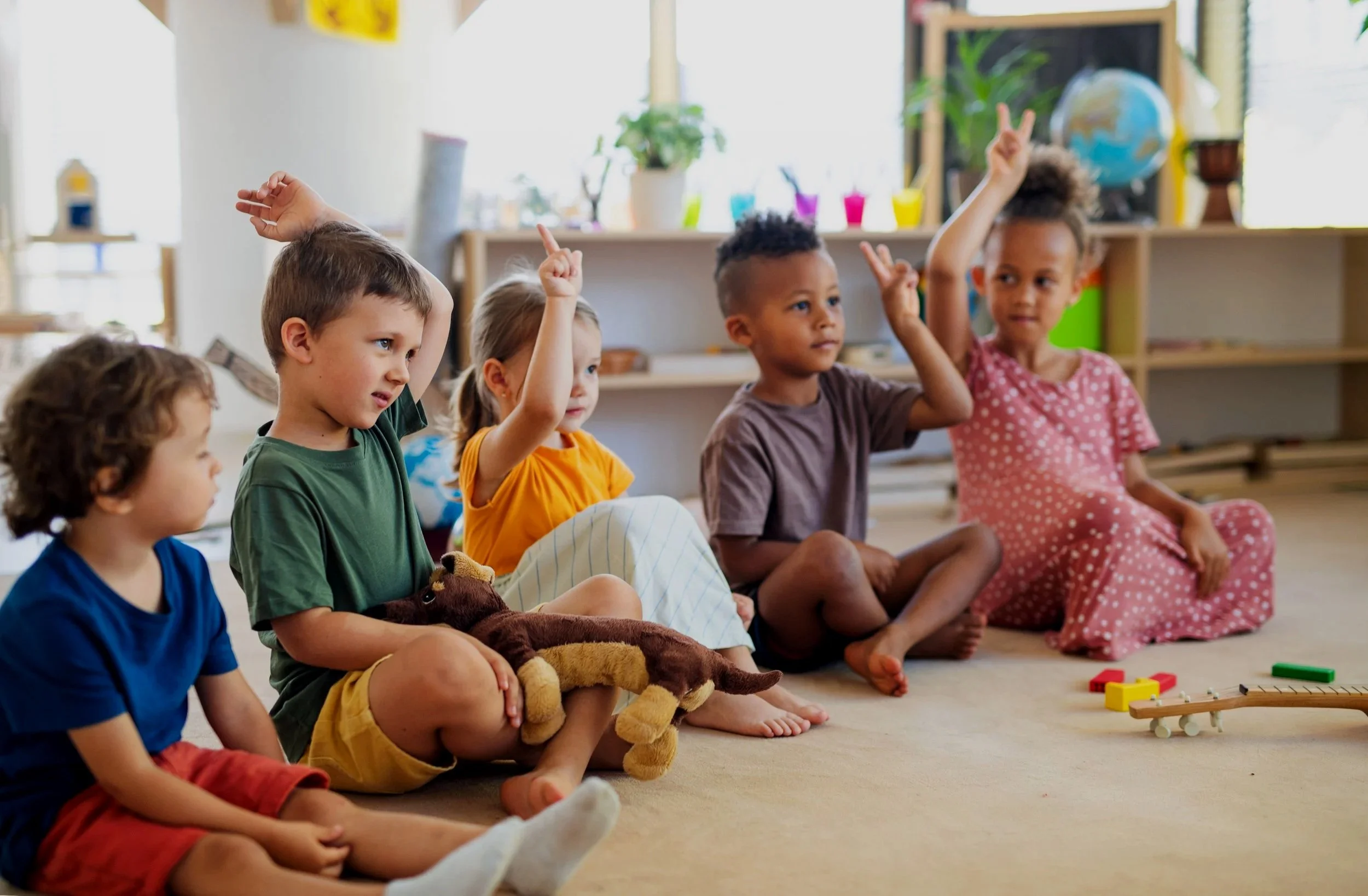 Group of five young children sitting on the floor in a classroom, raising hands for participation. The classroom has toys, plants, and educational decorations in the background.