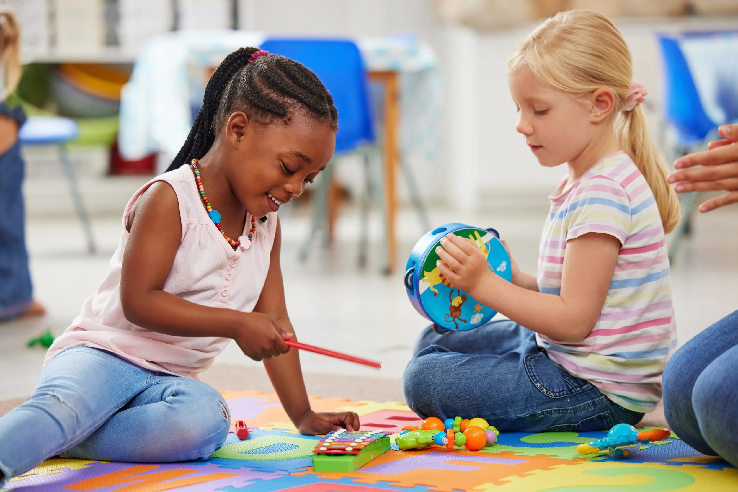 Two young girls sit on a colorful play mat playing with small toys, one girl with dark skin and braided hair wearing a pink sleeveless shirt and beaded necklace, the other with light skin and blonde hair in a ponytail wearing a striped shirt. They are smiling and interacting indoors.