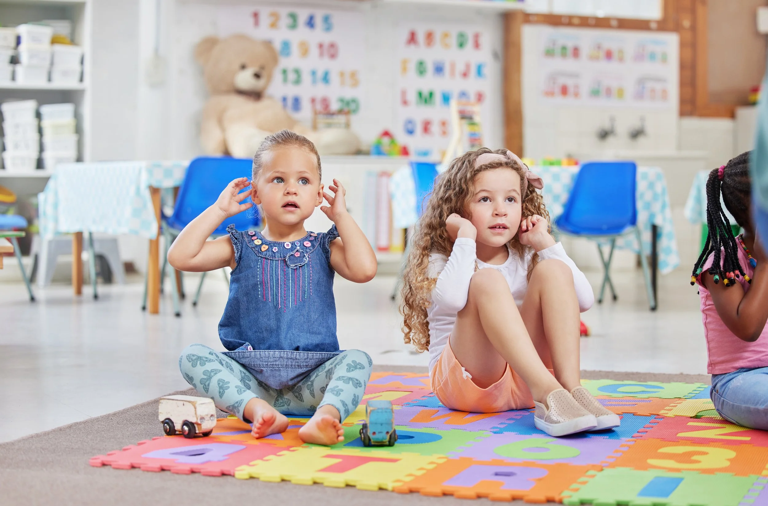 Two young girls sitting on a colorful foam play mat in a classroom. One girl has curly hair, white top, and orange shorts, the other has straight hair, a blue top, and butterfly leggings. Toys are on the mat. Classroom decorations include alphabet posters, numbers, and a large teddy bear.