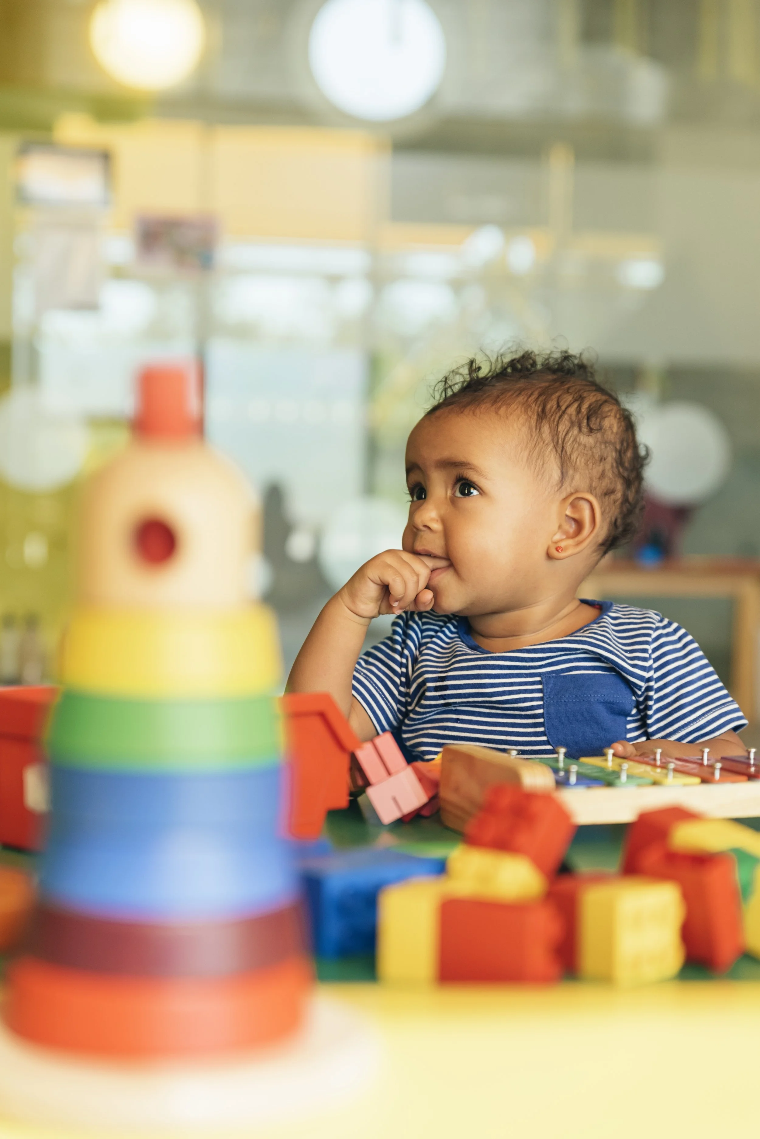 A young child with curly hair and a striped shirt sits at a table with colorful stacking blocks and games, focused and thoughtful.