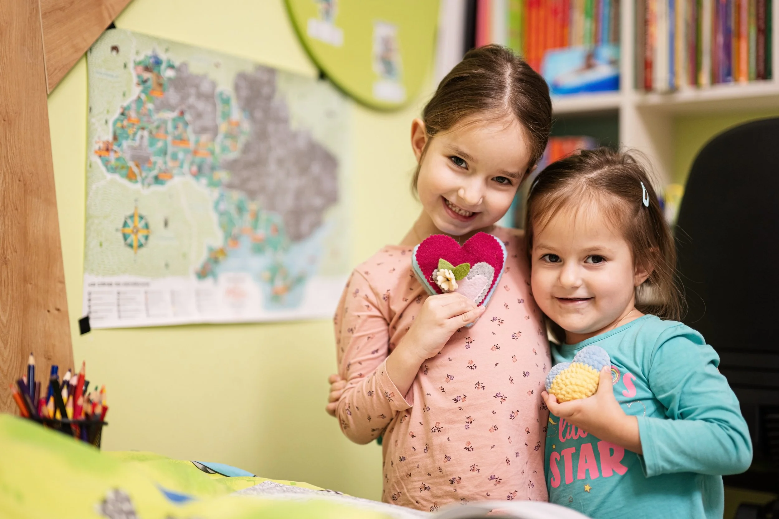 Two young girls standing close together, smiling, holding crafts, in front of a colorful wall and map, with art supplies on a table nearby.