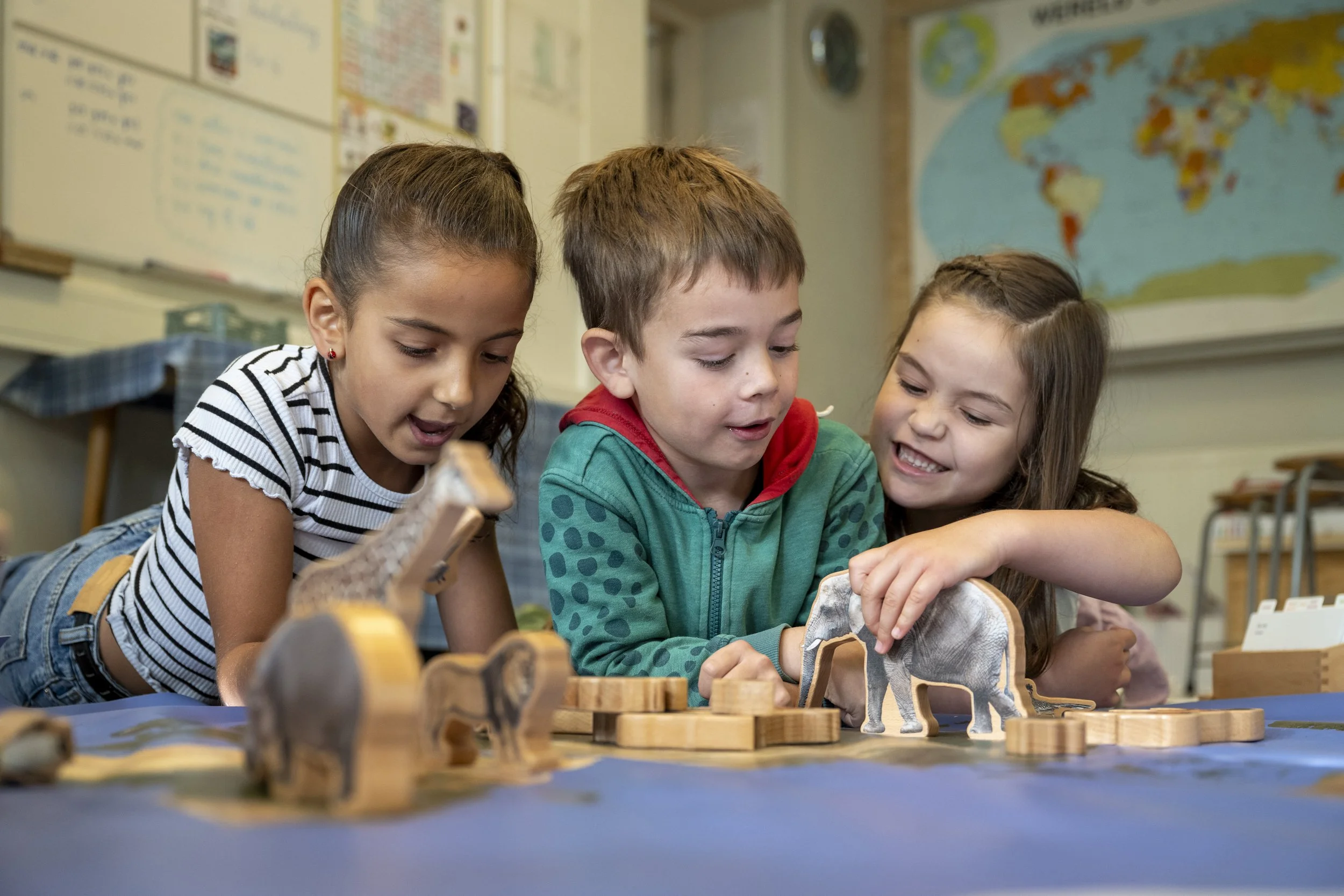 Three children, two girls and one boy, engaged in a tabletop game or activity involving miniature animal figures and wooden blocks in a classroom, with educational posters and a world map in the background.