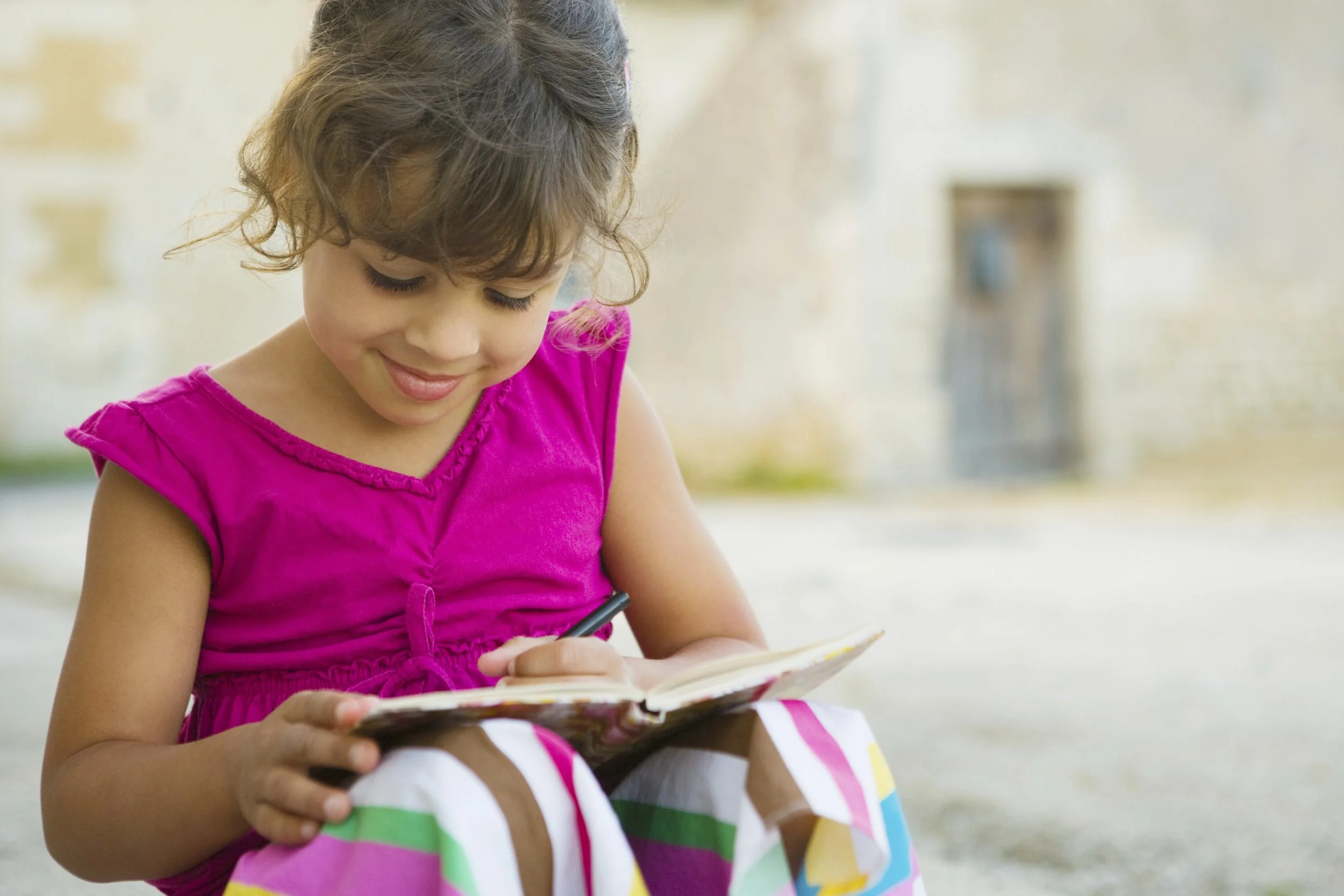 A young girl with curly hair wearing a pink top sitting outdoors, looking down and smiling at a colorful book she is holding.