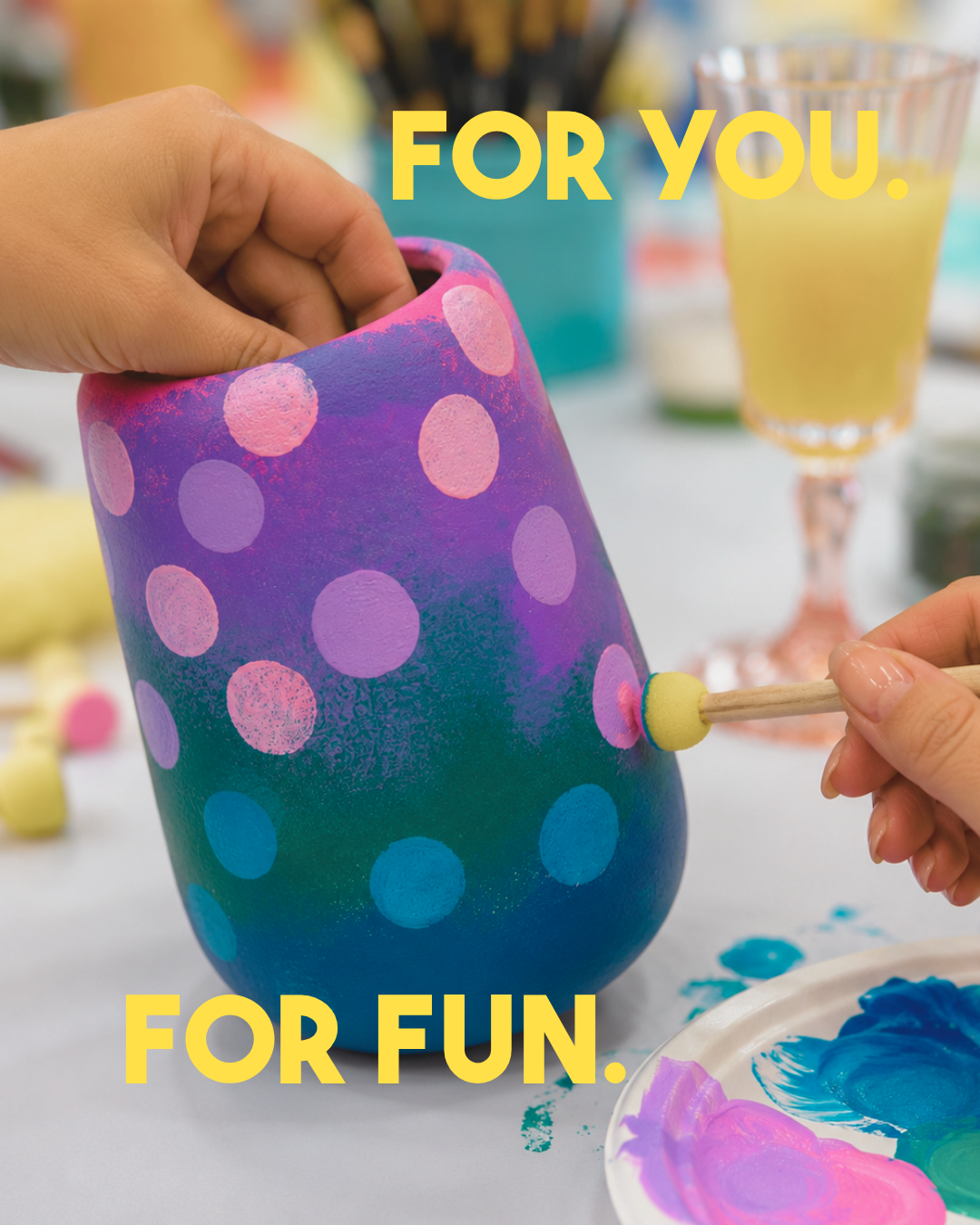 Person painting a colorful polka dot design on a ceramic vase with pink, purple, and blue dots, while another person paints a pink dot with a foam brush. The scene includes painting supplies and drinks in the background, with text "FOR YOU. FOR FUN."