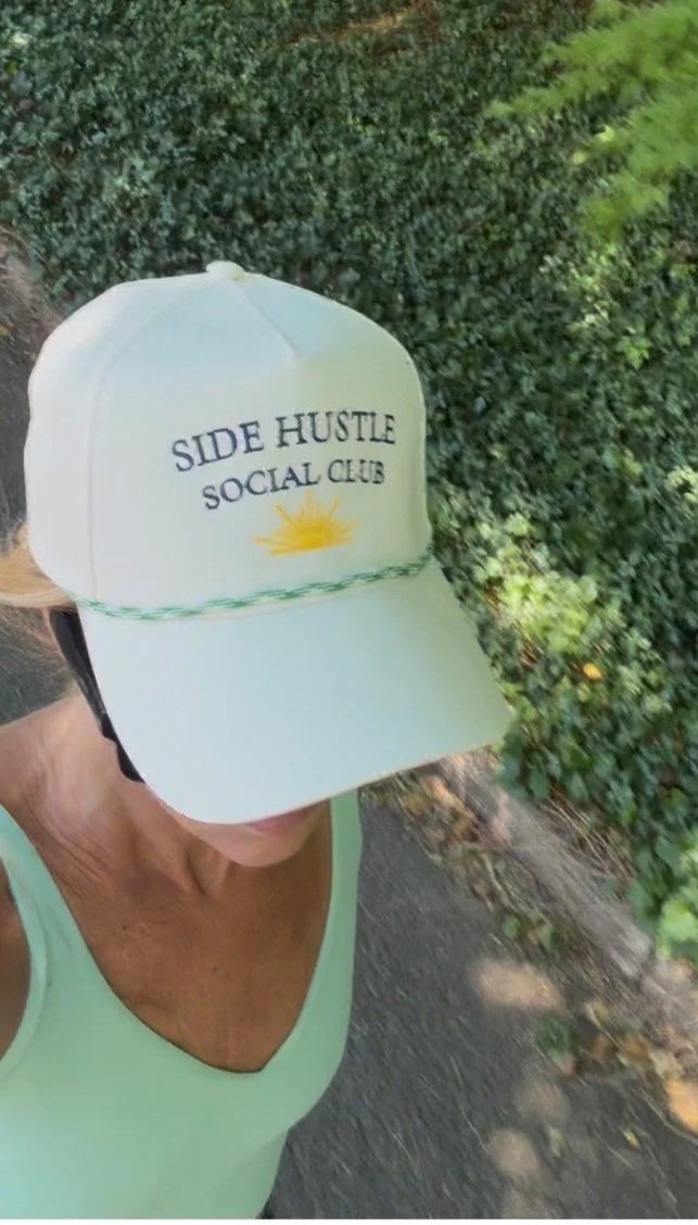 A person walking outdoors wearing a white baseball cap with the text 'Side Hustle Social Club' and a yellow sun graphic, a green tank top, and a colorful tattoo on their left arm, with green foliage and a pathway in the background.