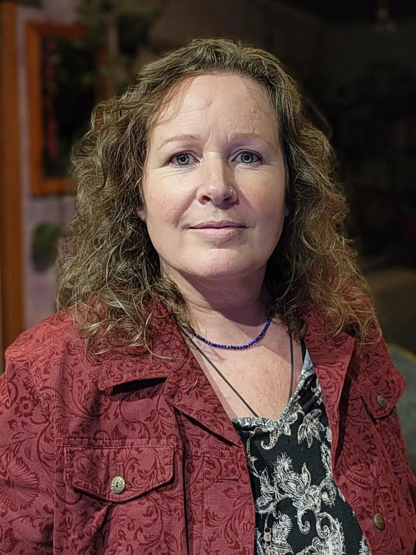 A woman with curly brown hair and blue eyes, wearing a black and white patterned shirt, a red jacket with floral designs, a blue necklace, and a black necklace, posing indoors with a dark background.