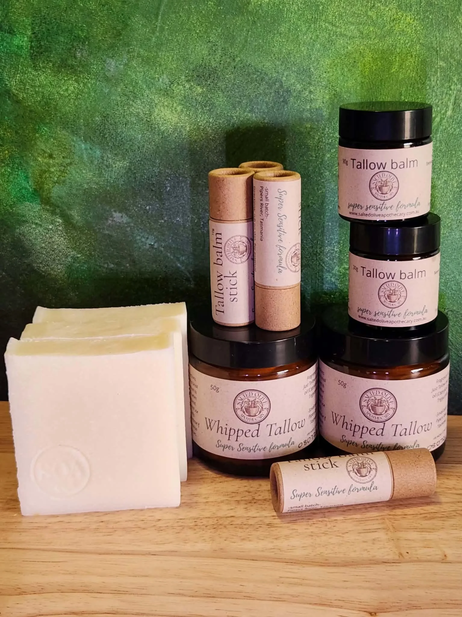 Various natural skincare products including whipped tallow, tallow balm, patchouli and vetiver sticks, and soap bars, displayed on a wooden surface with a green textured background.
