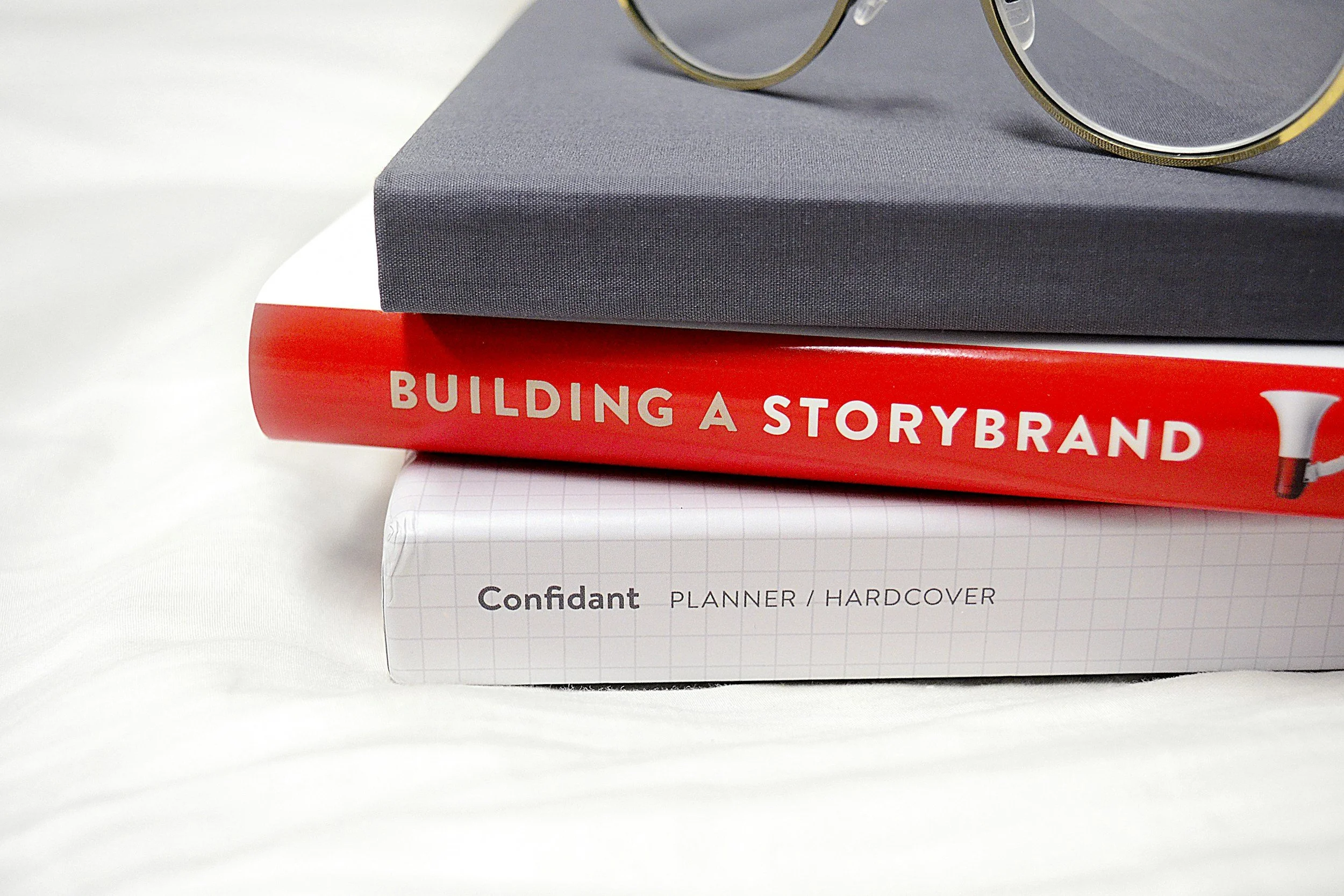 Three stacked books, with a gray hardcover book on top, a red book titled 'Building a StoryBrand' in the middle, and a white planner or notebook labeled 'Confidant Planner / Hardcover' at the bottom. Part of a pair of glasses is visible on top of the gray book.