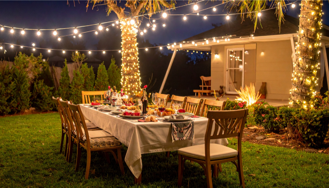 Outdoor evening dinner party in a backyard with a long dining table set with food, drinks, and tableware. Twinkling string lights are hung across two trees wrapped in fairy lights, creating a warm glow. In the background, there is a small house or shed with a bench, amidst a garden with shrubs and trees.