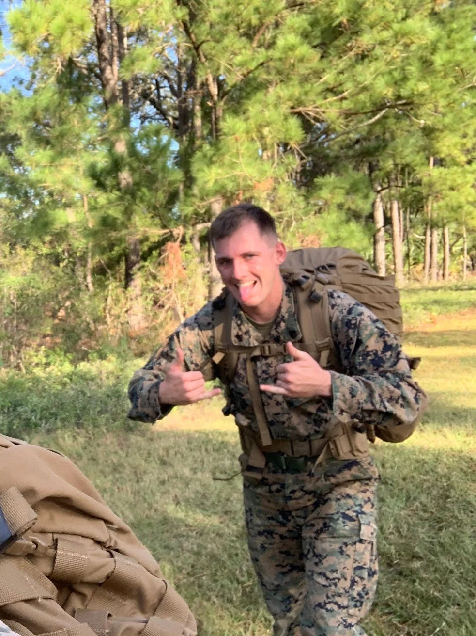 A soldier in camouflage uniform smiling and making a shaka sign with both hands in a forested area.