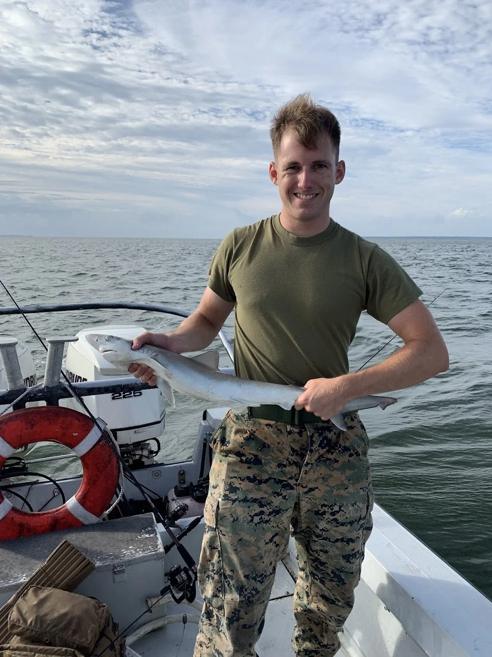 A young man in military camouflage pants and an olive green t-shirt on a boat holding a fish with a cloudy sky and open water in the background.