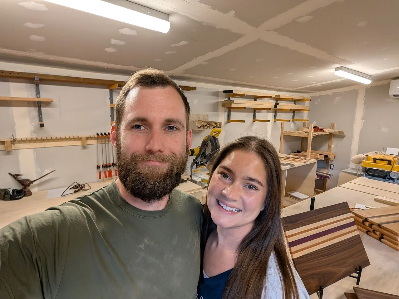 A smiling man and woman taking a selfie in a woodworking workshop. The workshop has shelves with woodworking tools and supplies, a workbench, and pieces of wood and finished wood projects.