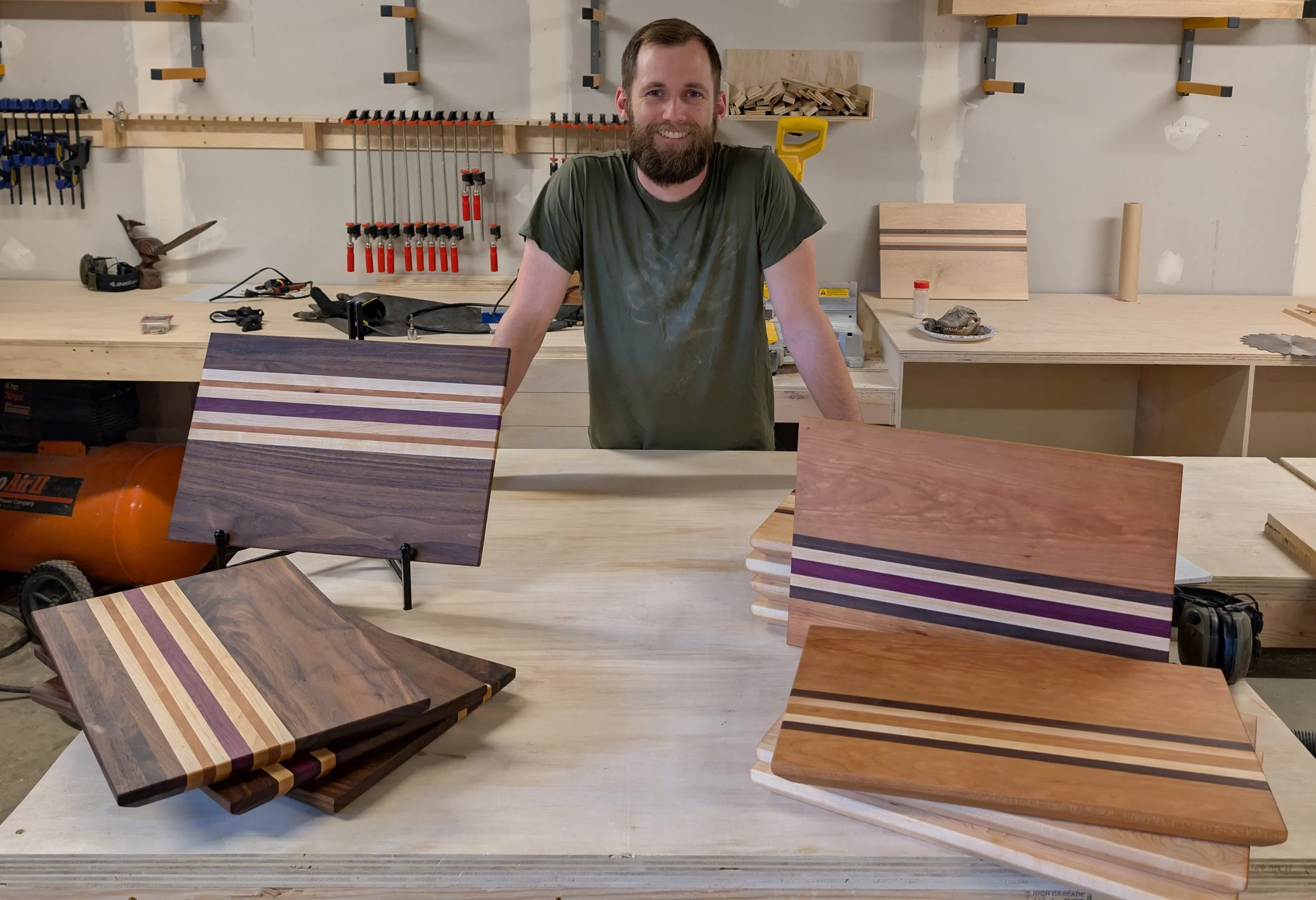 A man with a beard smiling stands behind a worktable in a woodworking workshop with various wood samples and tools arranged around him.