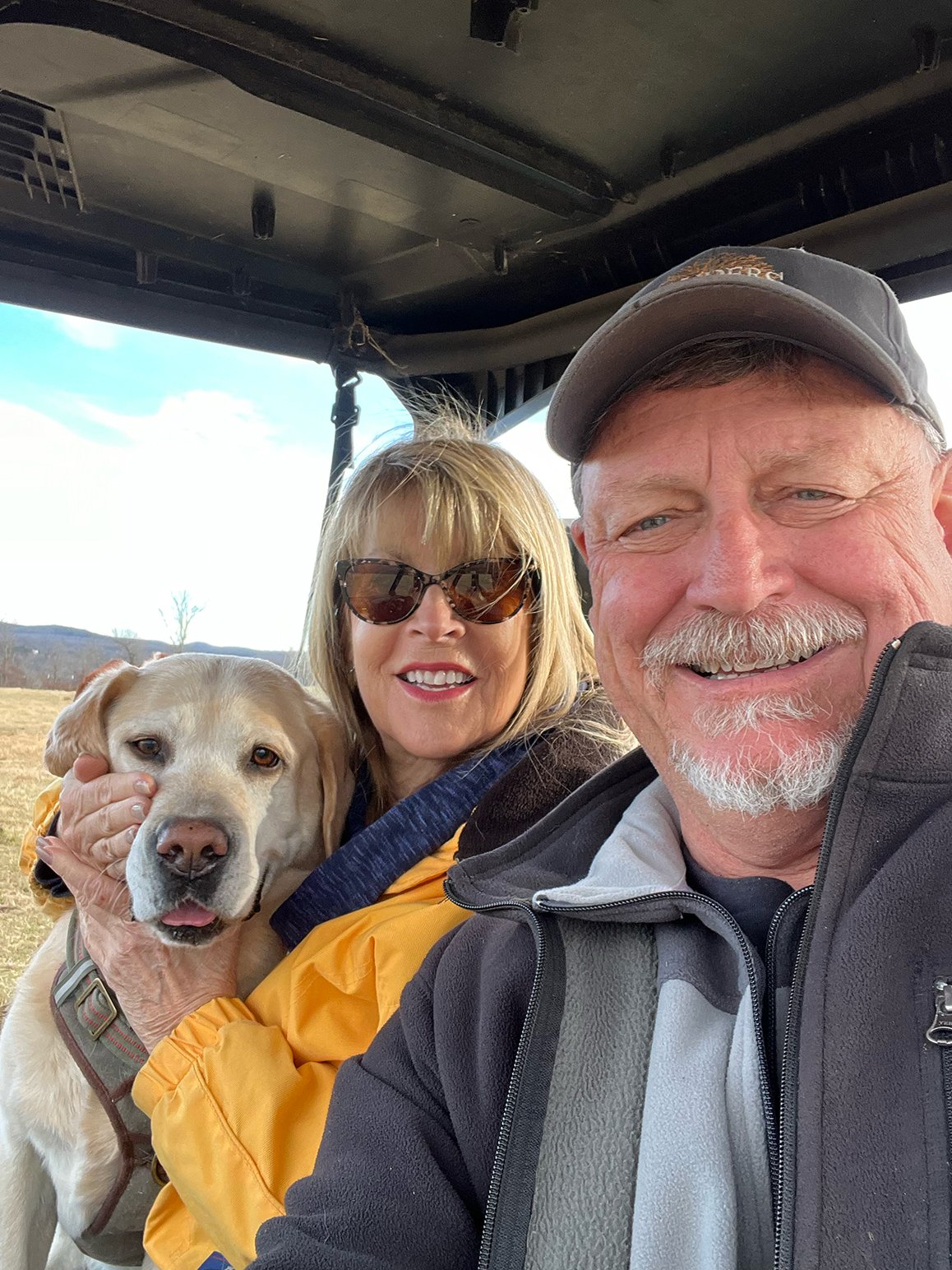 A smiling older man with a gray beard and a gray cap, a woman with blonde hair and sunglasses, and a Labrador Retriever dog, all sitting together outdoors on a sunny day.