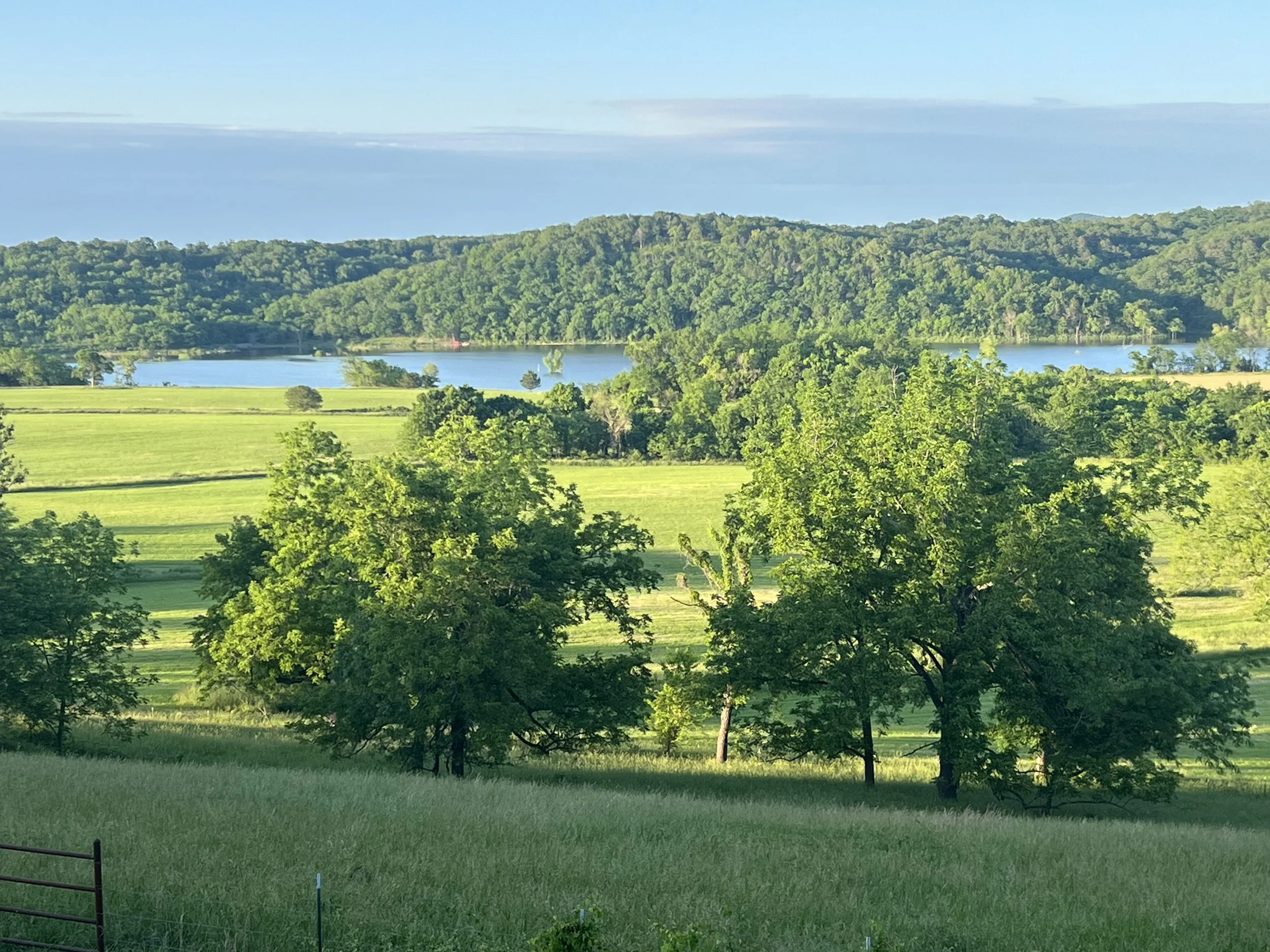 A landscape featuring green trees, rolling hills, and a lake under a partly cloudy sky.