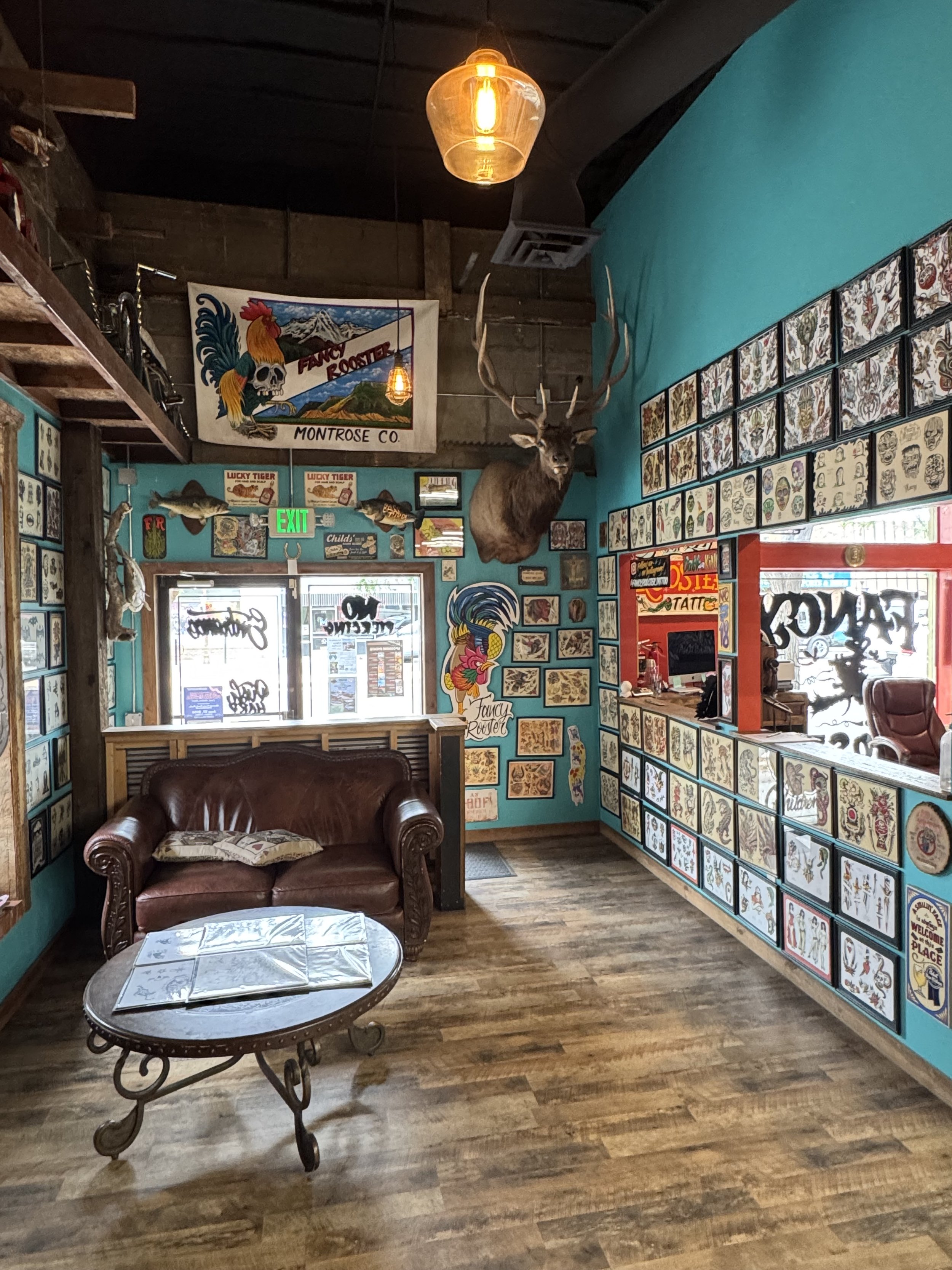 A cozy tattoo shop interior with light blue walls covered in framed tattoo art. There is a brown leather couch, a round coffee table with animal sketches, and a wall-mounted elk head with large antlers. A staircase with vintage bikes above leads to an upper level. Hanging light bulbs provide warm lighting, and a large trout fish is mounted on the wall near the entrance.