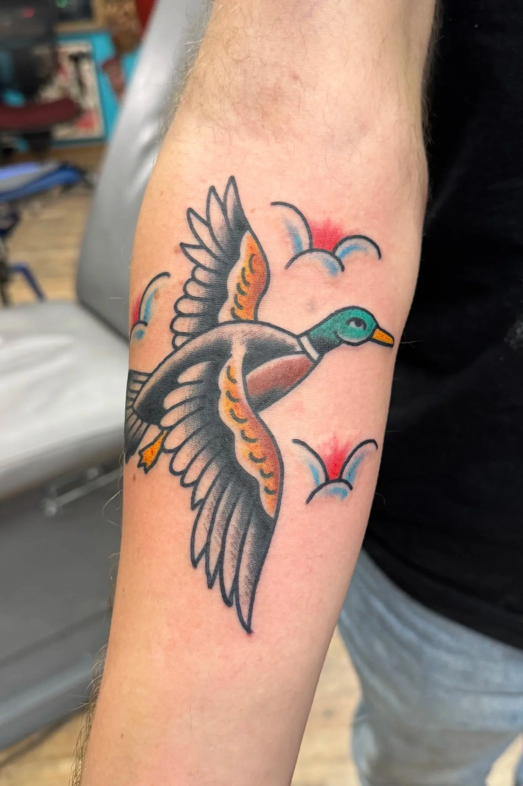 Colorful tattoo of a duck with a green head, orange beak, and stylized wings, set against a background of stylized clouds with red and blue hues.