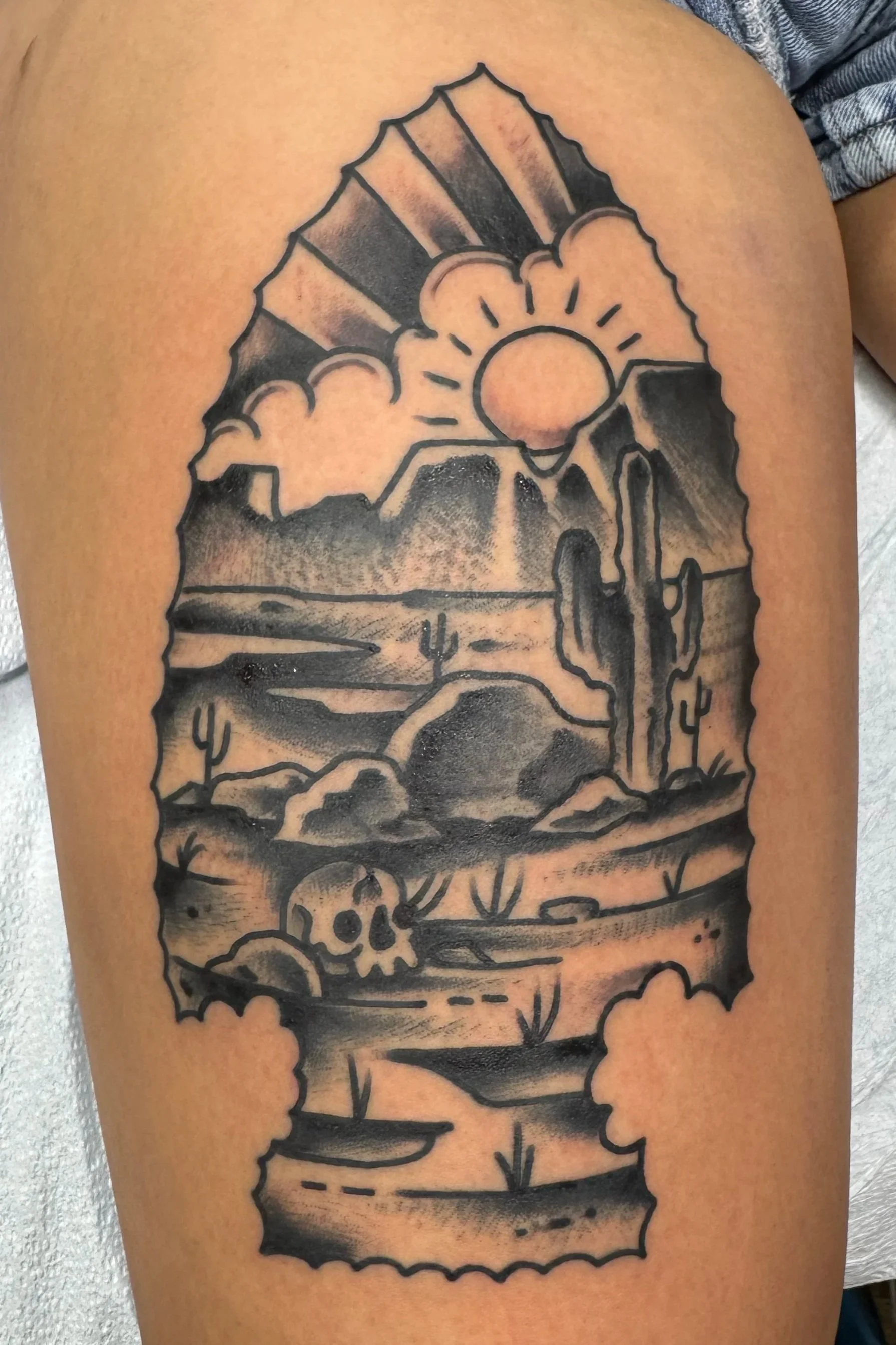 A black and gray tattoo of a portion of a colorado landscape on a person's upper leg, featuring a desert landscape with mountains, cacti, a setting sun, clouds, a skull, and various desert symbols.