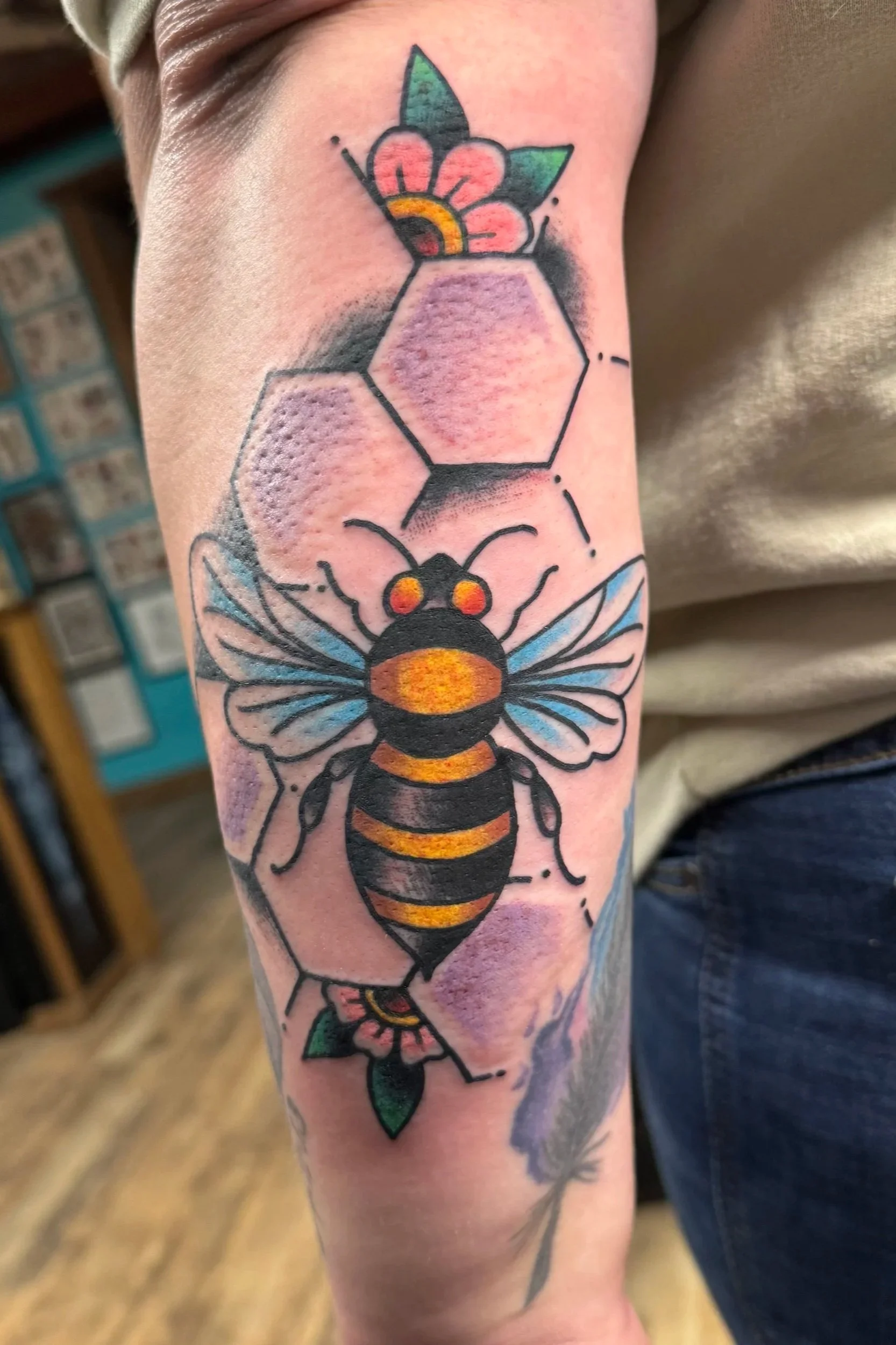Colorful tattoo on person's arm depicting a bee, honeycomb, and a pink flower with green leaves.