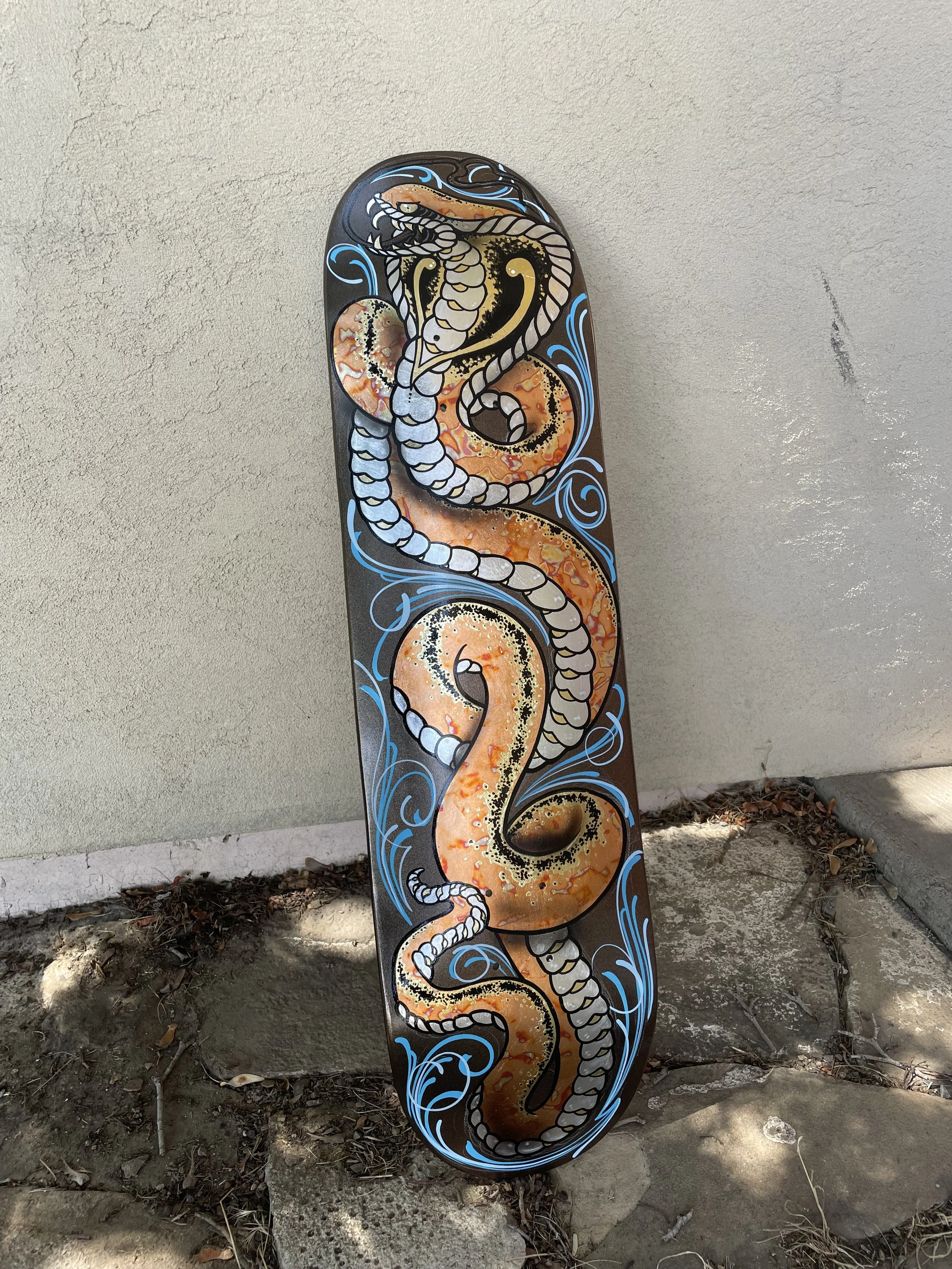 A skateboard with a colorful dragon design, featuring a long, winding body with white scales and blue accents, is leaning against a beige wall outdoors on a concrete and dirt surface.