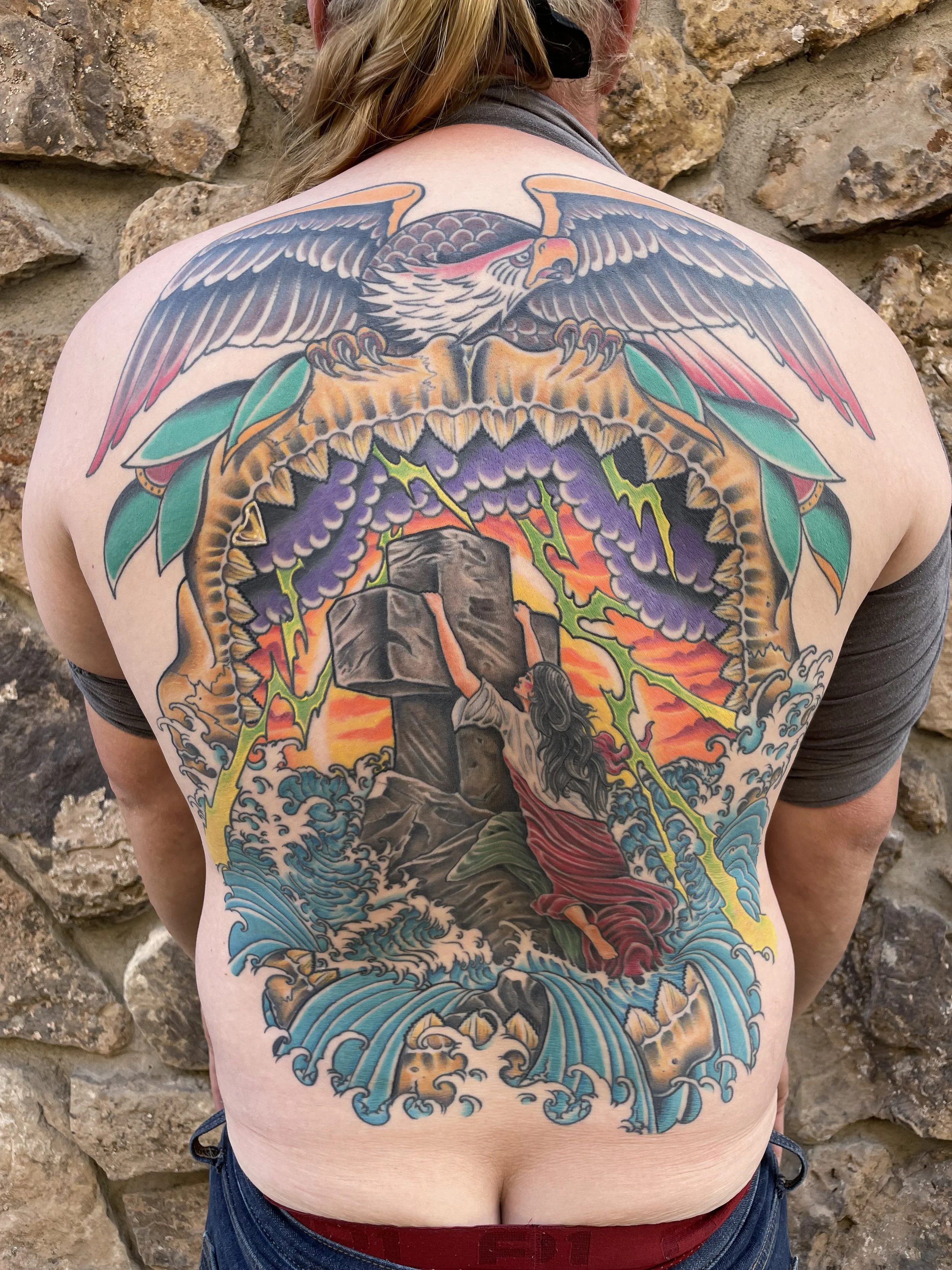 Back tattoo of an eagle with detailed feathers, colorful background with waves, and a woman holding a torch.