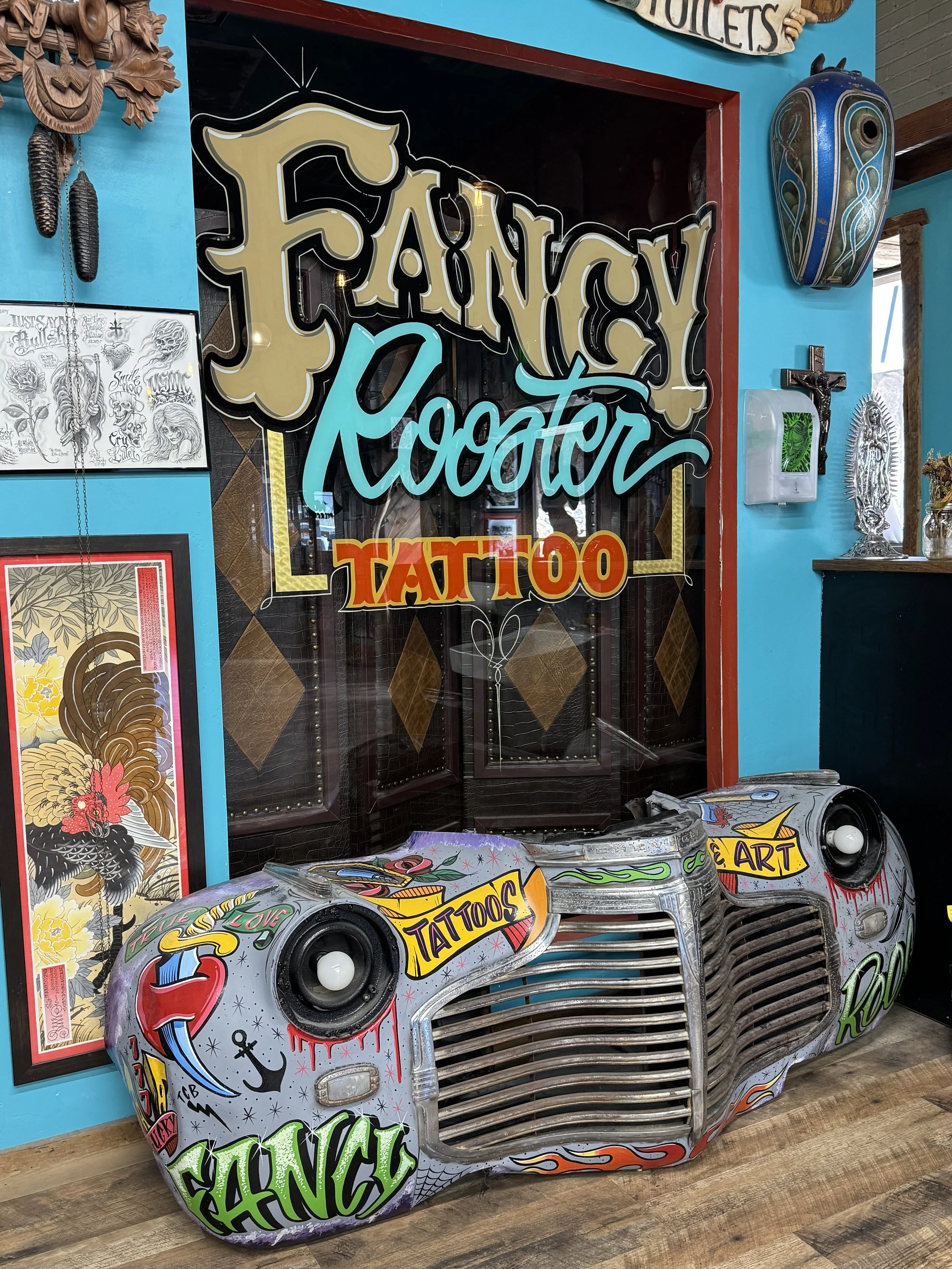 Decorative truck hood with tattoos, graffiti-style art, and words like 'Dance' and 'Love' painted on it, placed on a wooden floor inside a colorful tattoo shop.