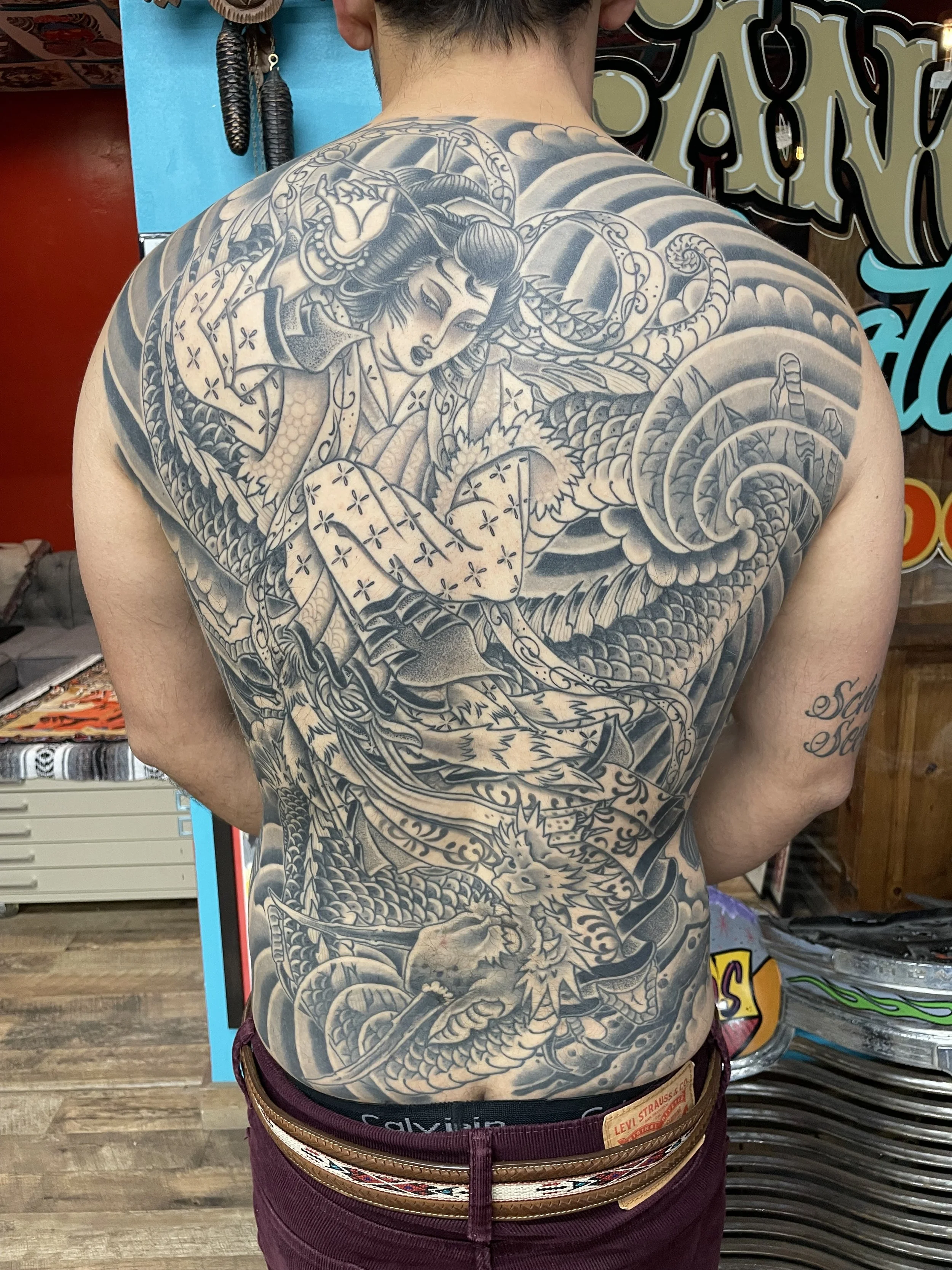 Back view of person with a detailed black and gray Japanese-style sleeve tattoo on the back and a dragon tattoo on the lower back, standing in a tattoo shop.