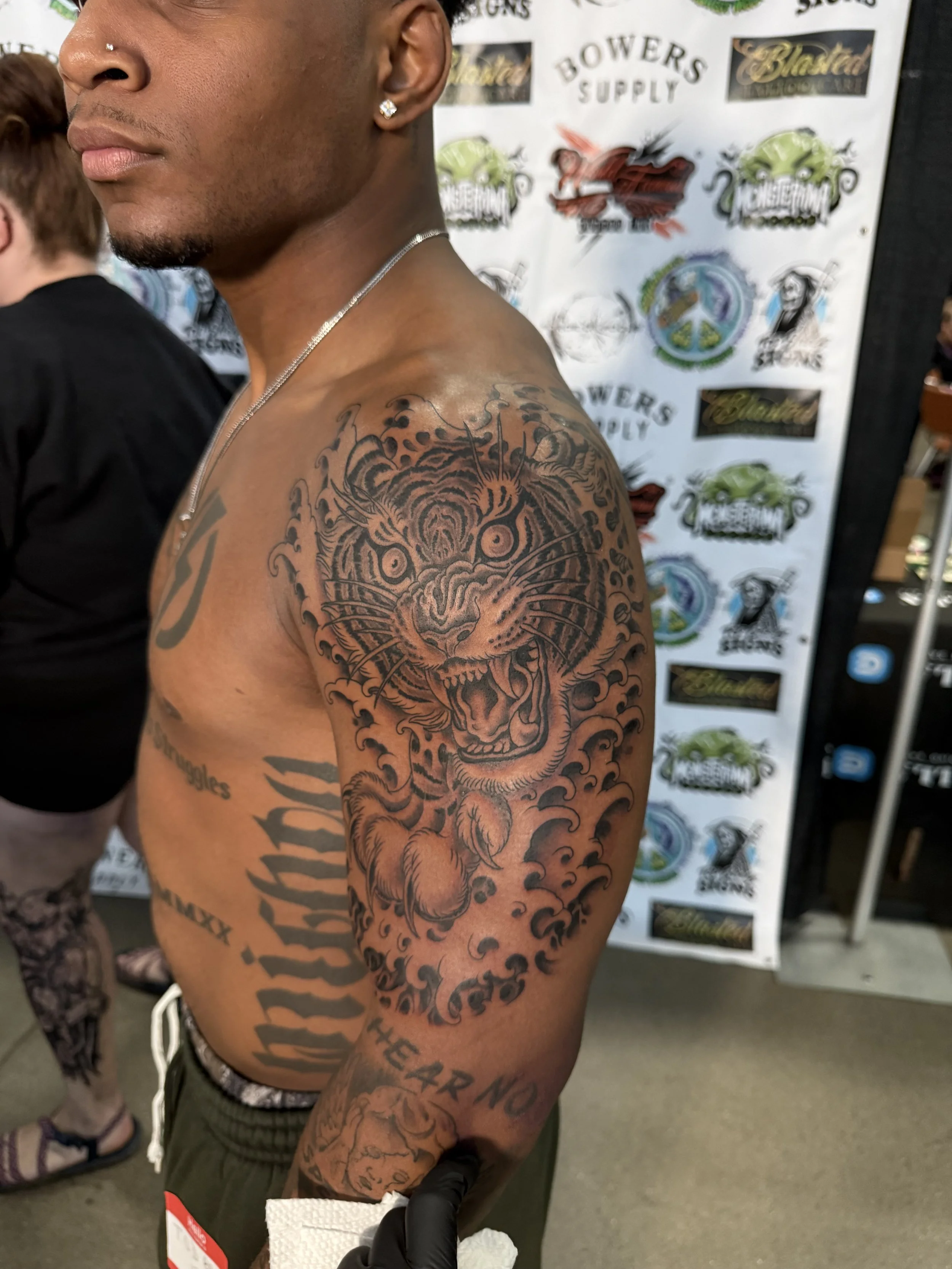 Close-up of a shirtless man with a large tattoo of a fierce tiger on his left shoulder and upper arm, wearing a necklace, earrings, and a nose piercing, standing in front of a backdrop with logos and designs, holding a shopping basket.