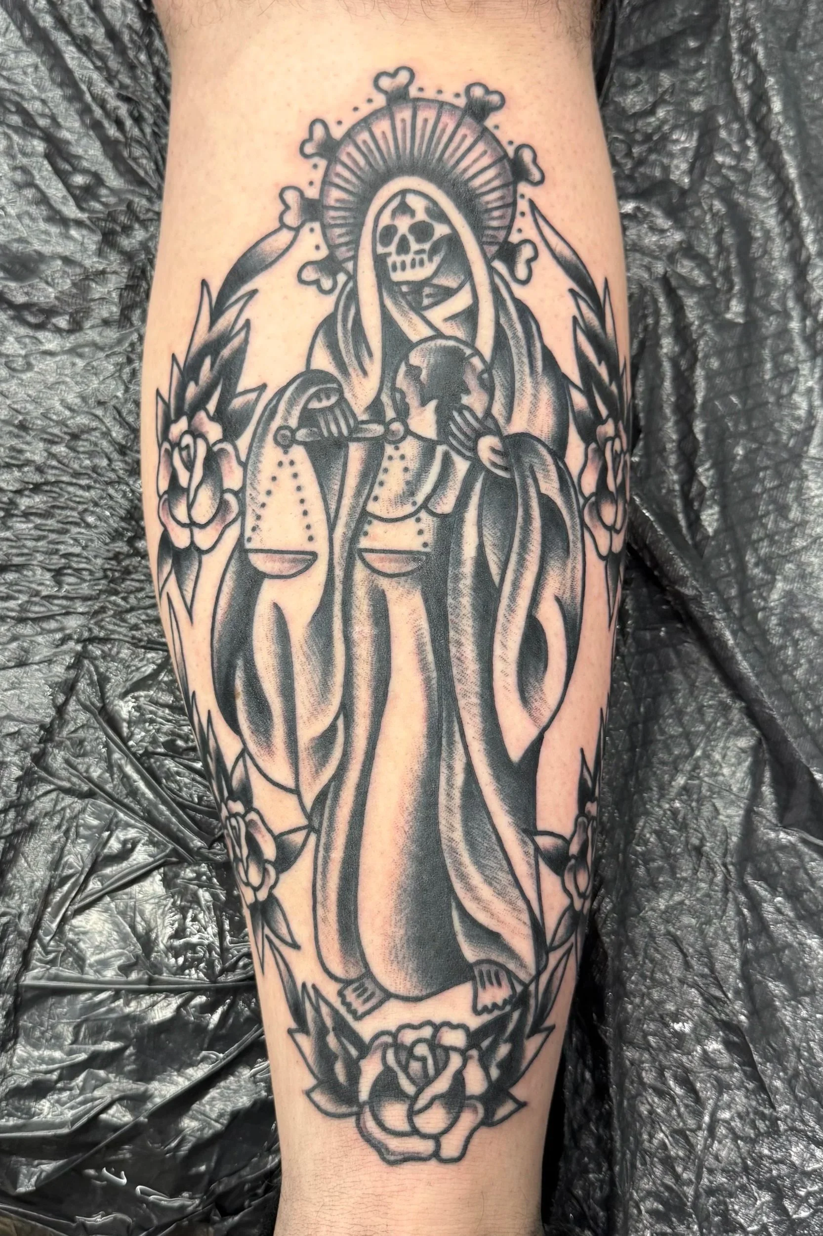 Tattoo of a skeletal figure wearing a hooded robe, holding a globe, with a halo and surrounded by roses on a person's arm.