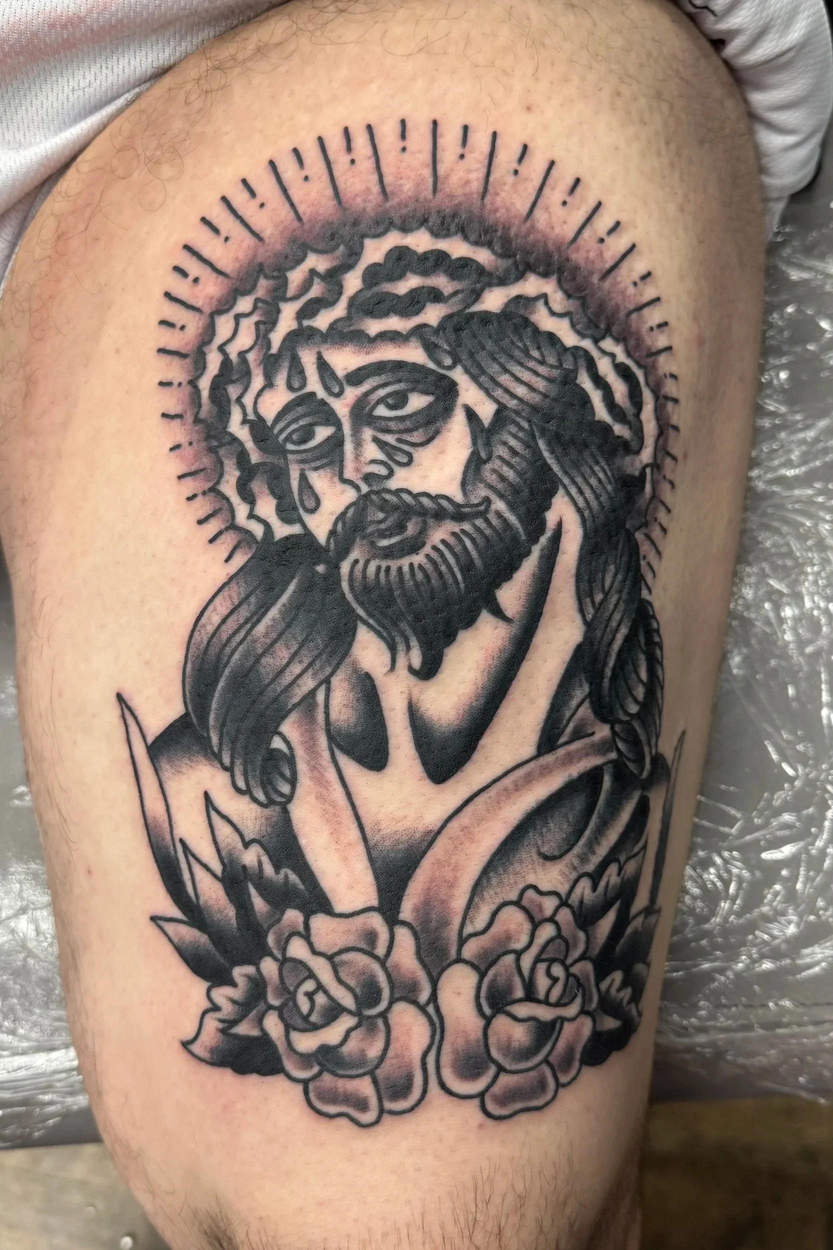 A black and gray tattoo of Jesus Christ with blood and tears on his face, surrounded by a halo of lines, on a person's upper arm.
