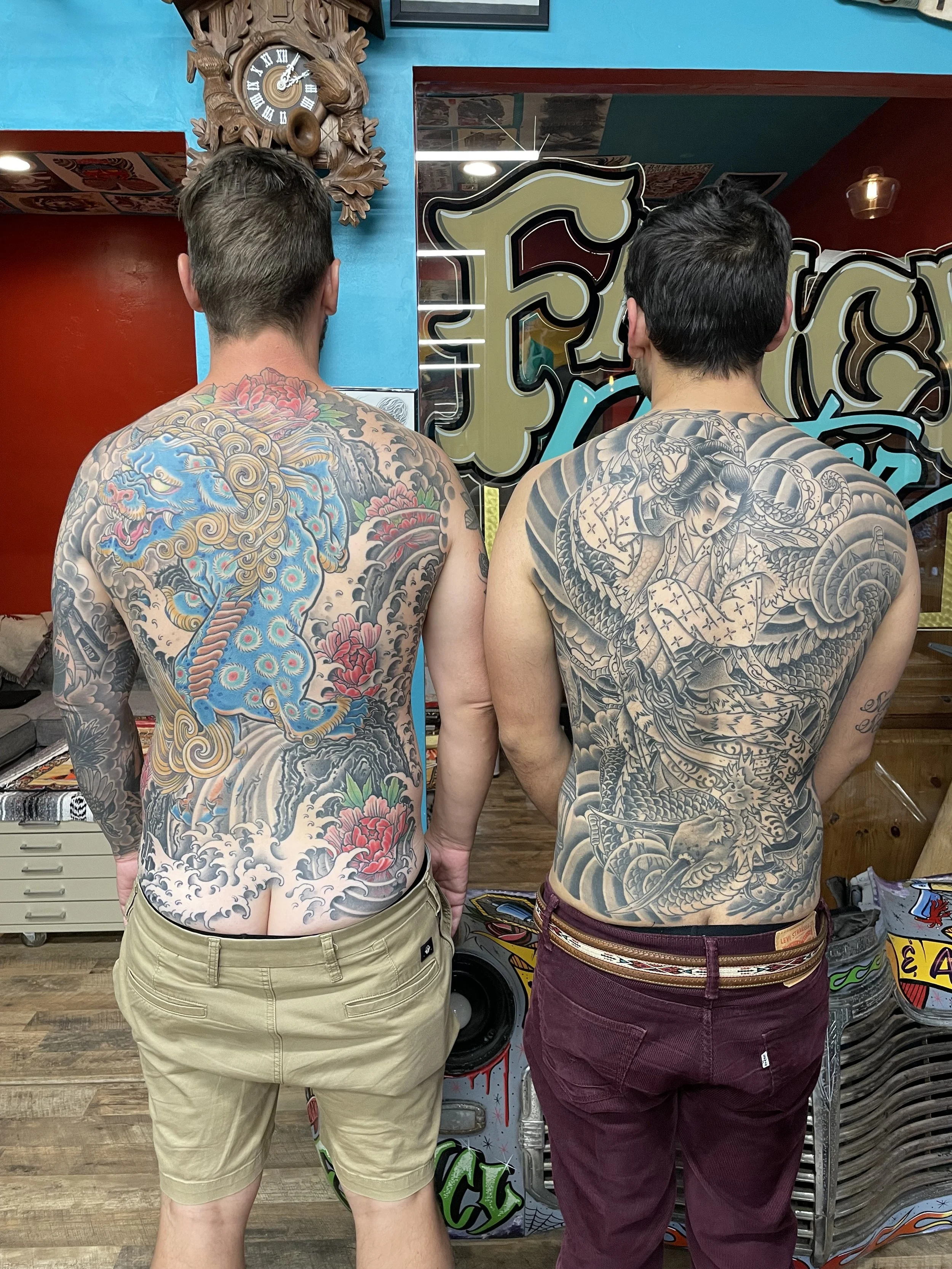 Two men with tattoos on their backs standing indoors, facing away from the camera. The man on the left has a colorful Japanese dragon tattoo, wearing khaki shorts. The man on the right has a black and gray traditional Japanese style tattoo, wearing maroon pants. Behind them, there is a red wall, a light blue wall, and decorative elements including a wooden wall clock and vintage signs.